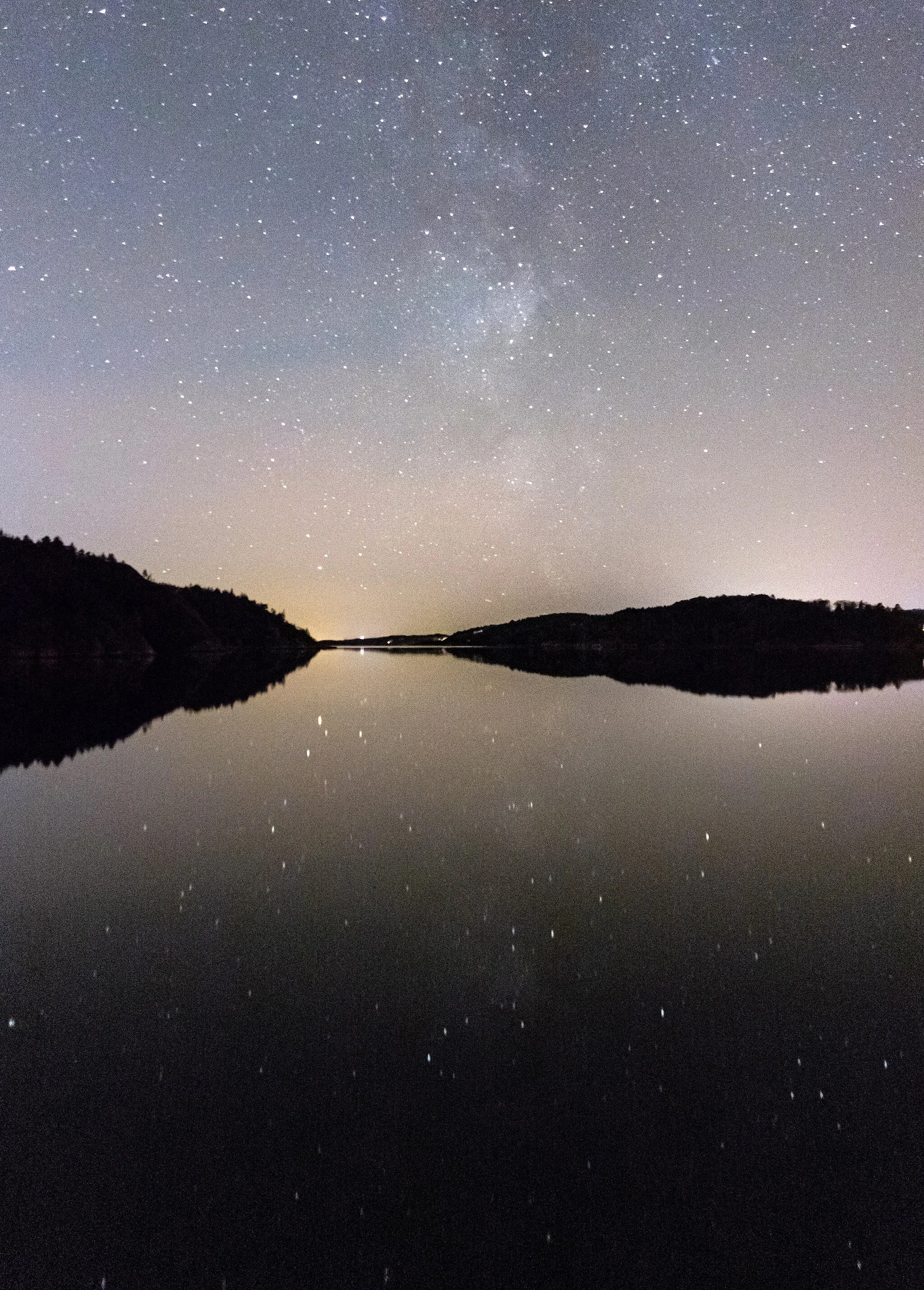 Night sky filled with stars and the Milky Way over a calm lake reflecting the starry sky and silhouetted hills. As above, so below.