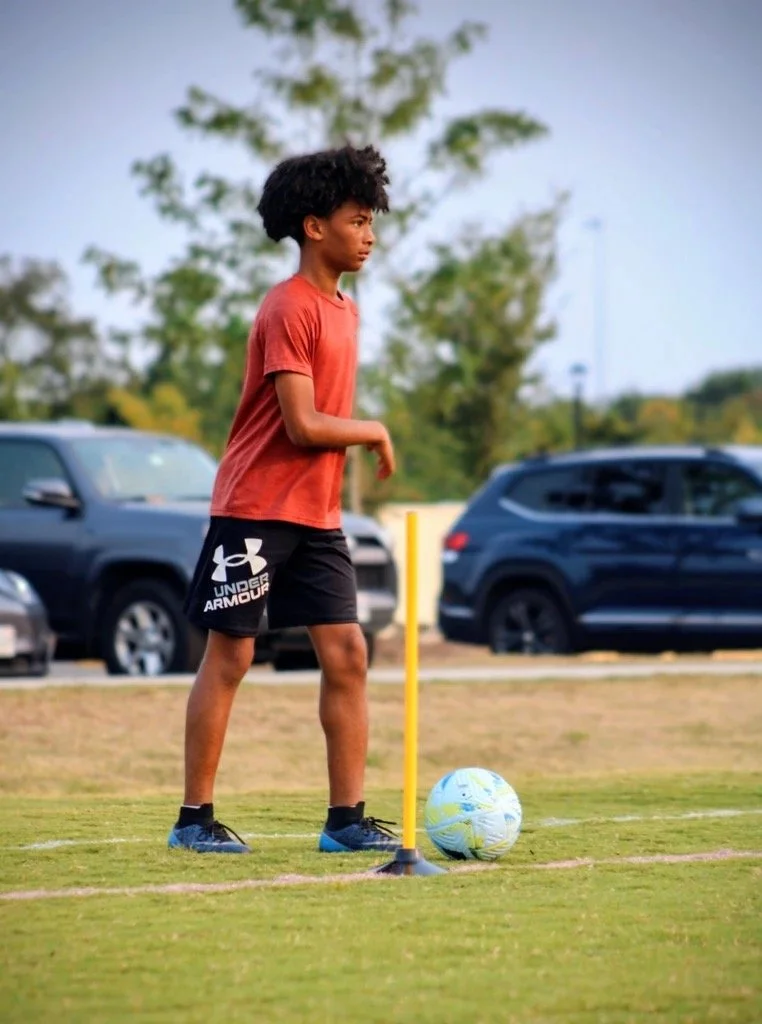 A young boy with black curly hair wearing a red T-shirt and black Under Armour shorts with a soccer ball, a yellow training pole, and playing field in the background.