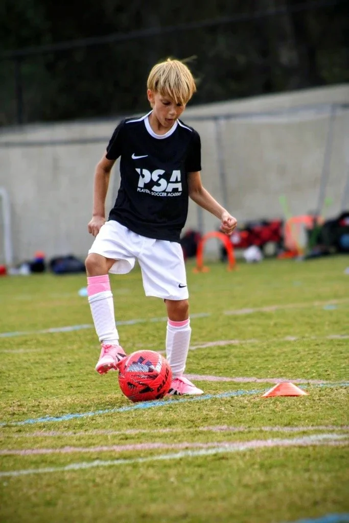 Young boy practicing soccer on a field, wearing a black jersey with 'PSA' logo, white shorts, and pink shoes, with a red and black soccer ball at his feet and orange cones set up for drills.