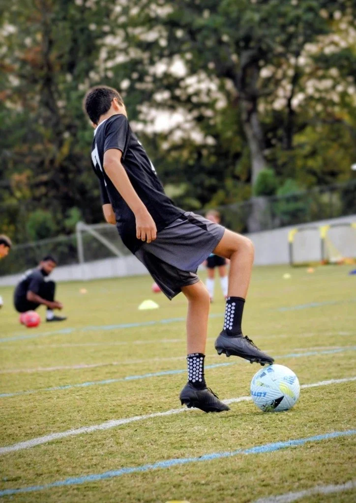 Boy in black sports uniform practicing soccer on a field with others in the background.