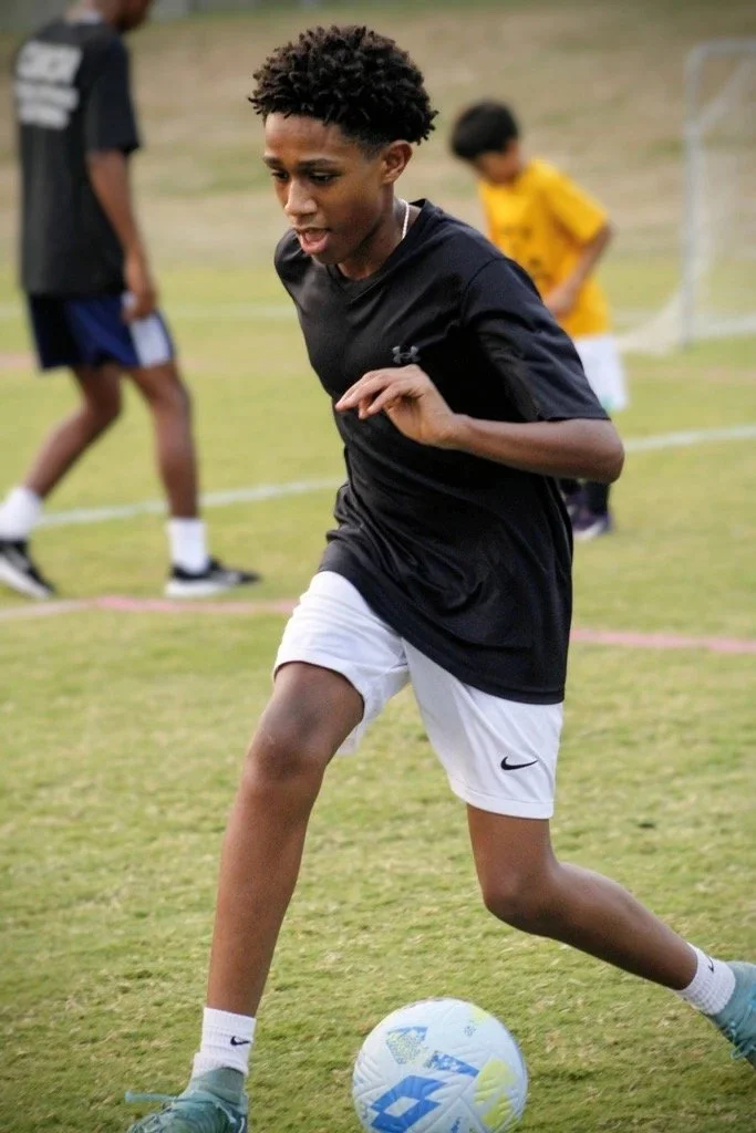 Young boy playing soccer outdoors on a grassy field, wearing a black shirt, white shorts, and blue shoes, dribbling a soccer ball.
