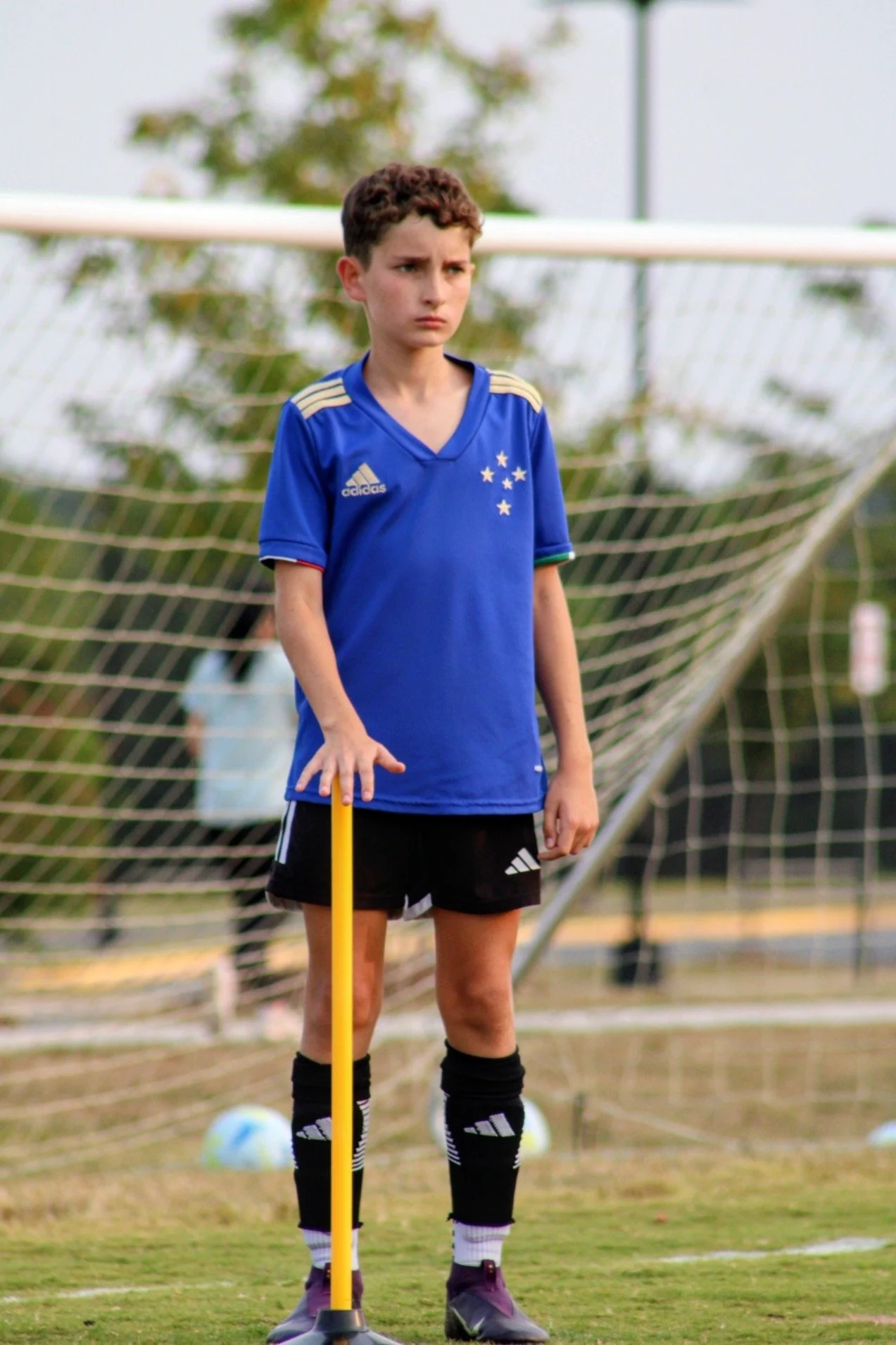 Young boy standing on a soccer field, dressed in a blue jersey and black shorts, holding a yellow training pole, with a soccer goal and blurred background behind him.