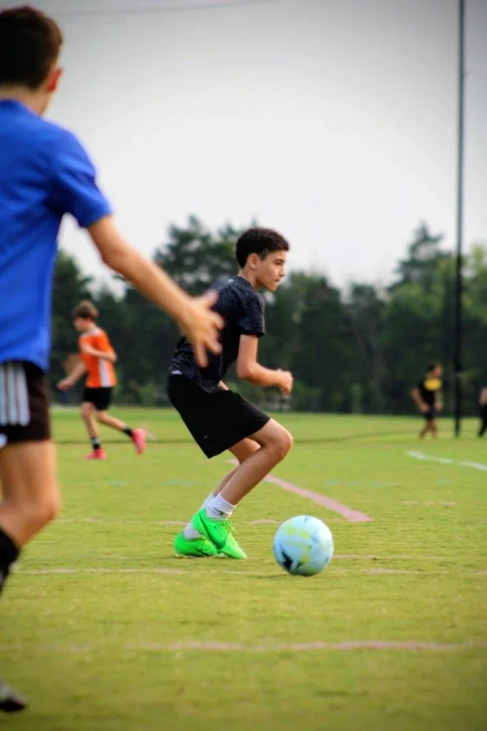A young boy in black shorts and neon green cleats playing soccer on a grassy field, with other players in the background.