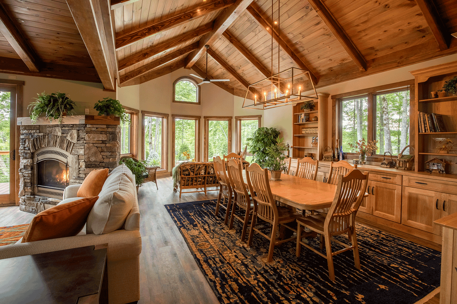 Living and dining area with wood ceiling, stone fireplace, large windows showing green trees, wooden dining table with eight chairs, built-in wooden shelves, plants, and a black patterned rug.