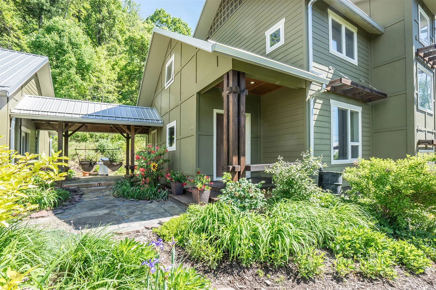 A modern green house with multiple windows and a small porch. There is a landscaped garden with various plants and flowers in front, and a covered outdoor sitting area with hanging chairs and hammocks in the background.