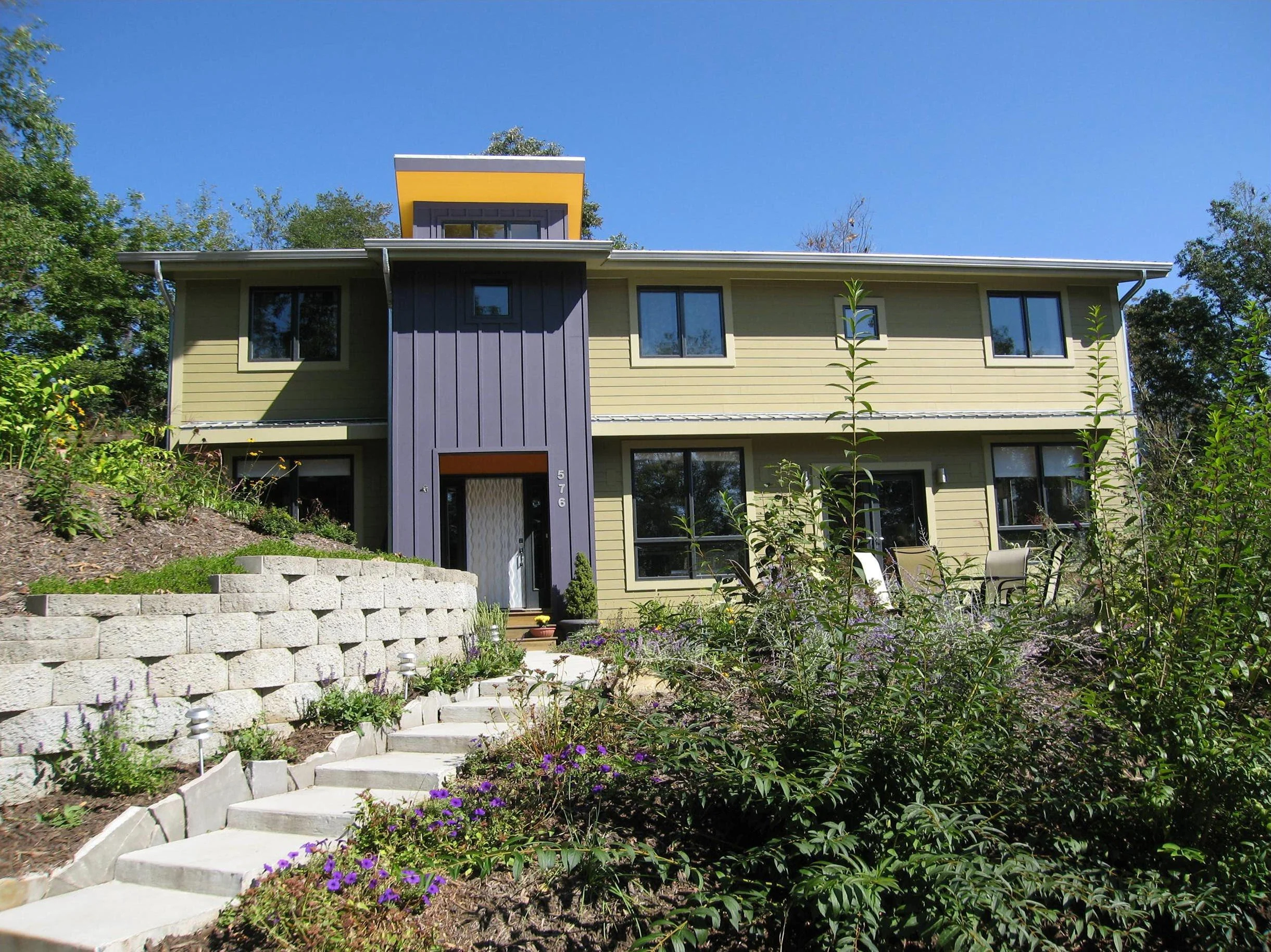 A two-story house with yellow siding, dark blue vertical accents, and large windows, with a landscaped front yard with purple flowers and a concrete staircase leading to the entrance.