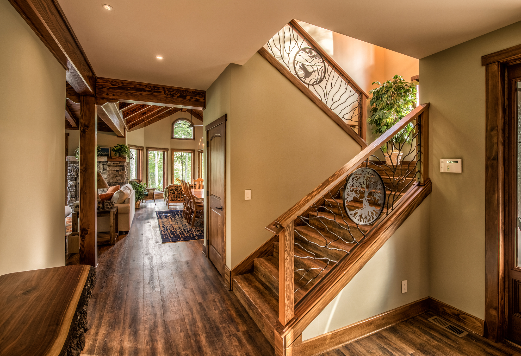 Interior of a home featuring a staircase with decorative metal railing, a wooden floor, and a view into a living and dining area with large windows, a stone fireplace, and wooden furniture.