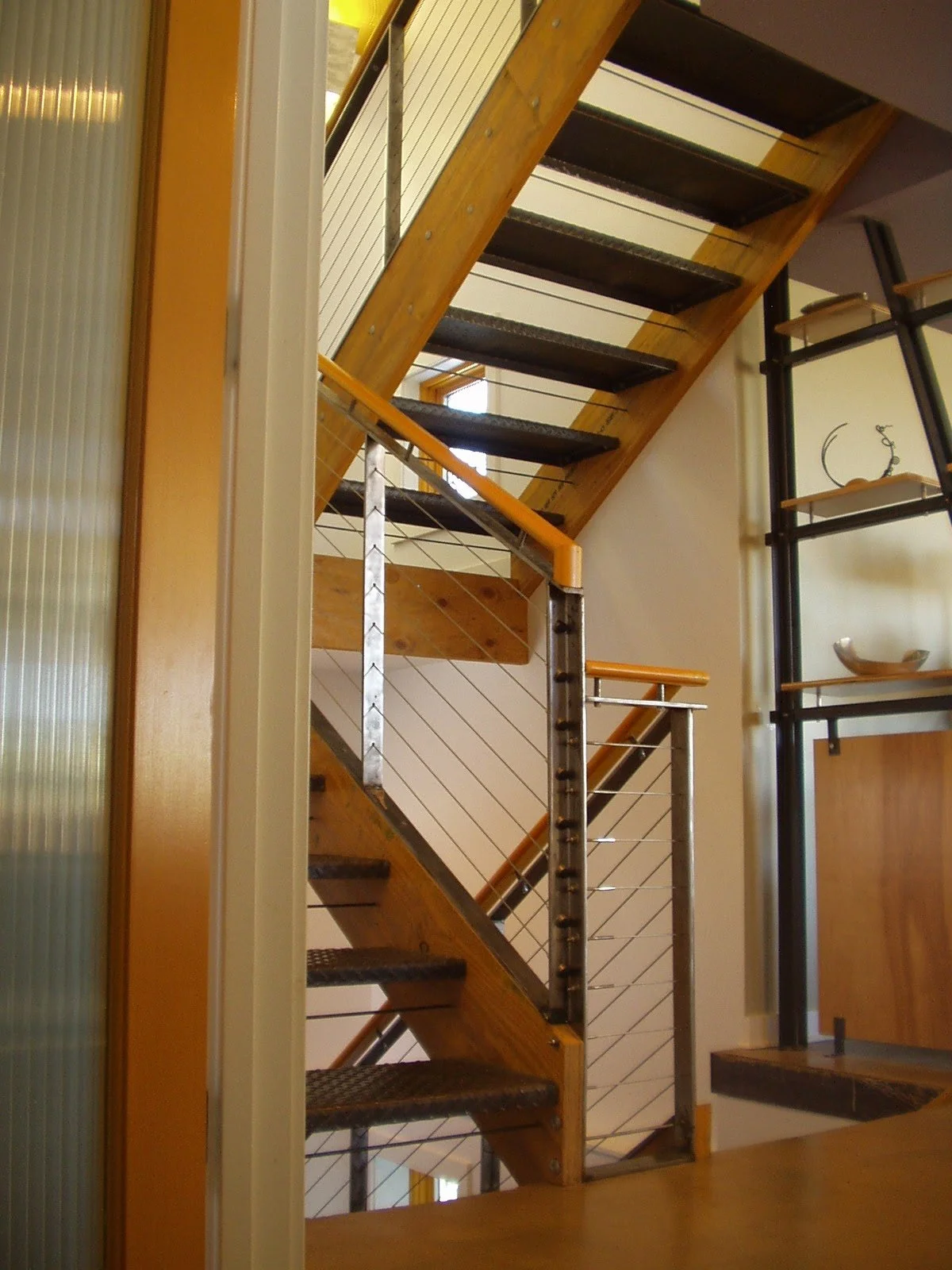 Interior view of a modern staircase with wooden steps, metal supports, and cable railing, leading upwards.