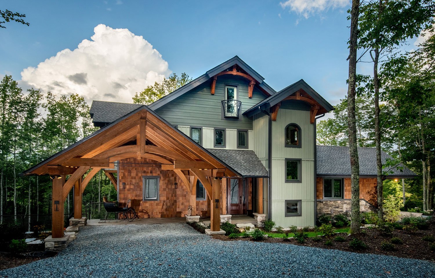A modern multi-story house with a combination of wood, stone, and vertical siding, featuring a gabled roof and a small balcony, surrounded by trees and a gravel driveway.