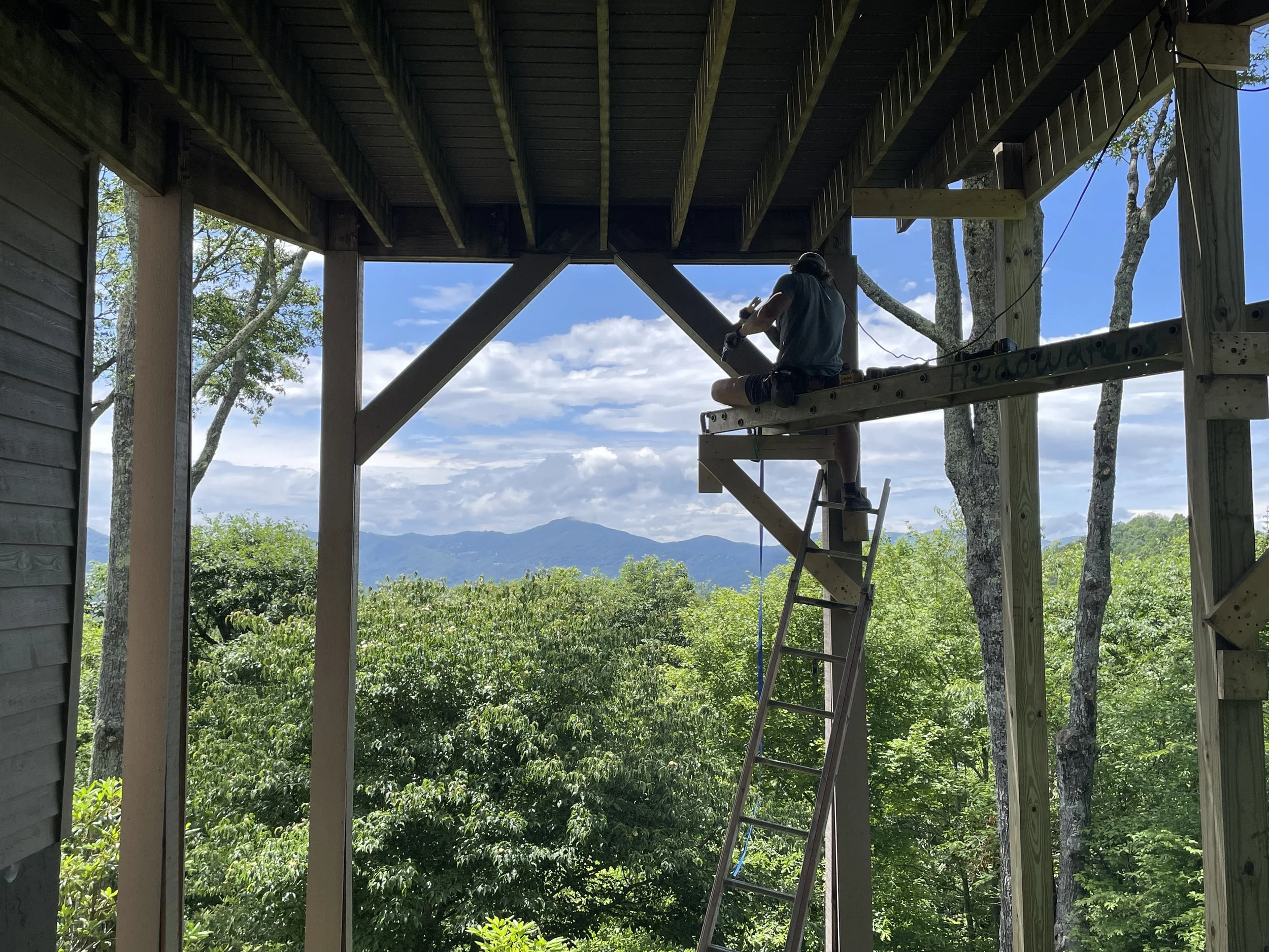 Person sitting on a wooden construction platform under a roof, working on a structure with tools, surrounded by trees and mountains in the background.