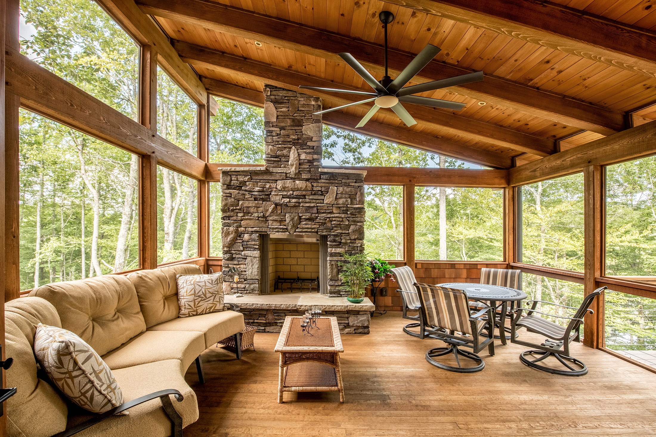 Sunroom with large windows, wood ceiling, stone fireplace, beige sofa with pillows, round dining table with striped chairs, ceiling fan, and kitchen counter with plants, overlooking a forest.