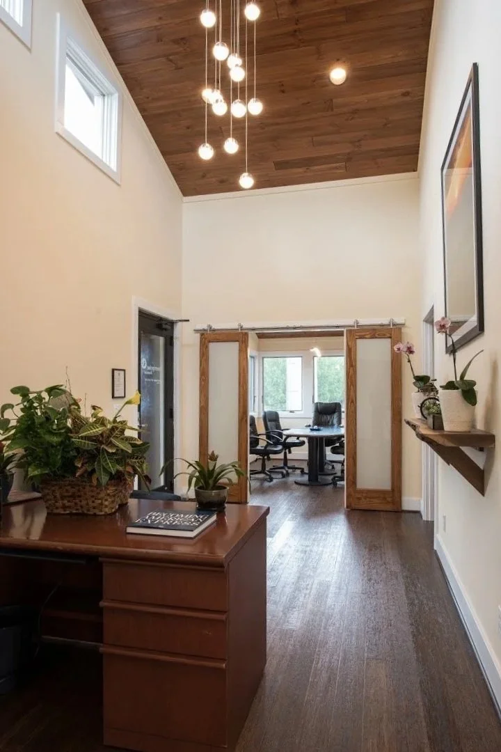 Interior view of a hallway leading to a conference room with large windows, a round table, and office chairs. The hallway has a wooden floor, cream walls, and a wooden ceiling with modern hanging light fixtures. There are houseplants on a desk and a wall shelf.
