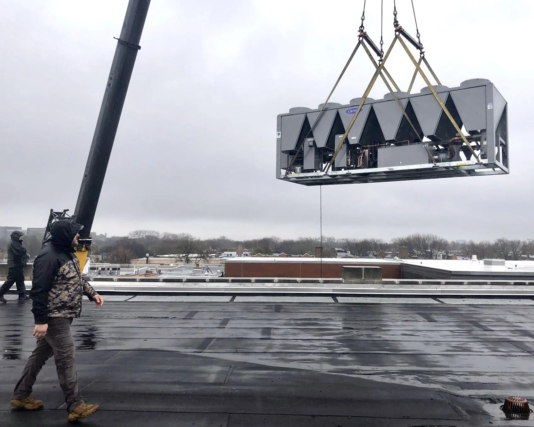 A person operating a crane on a rooftop, installing large HVAC units. The sky is overcast and the rooftop is wet.