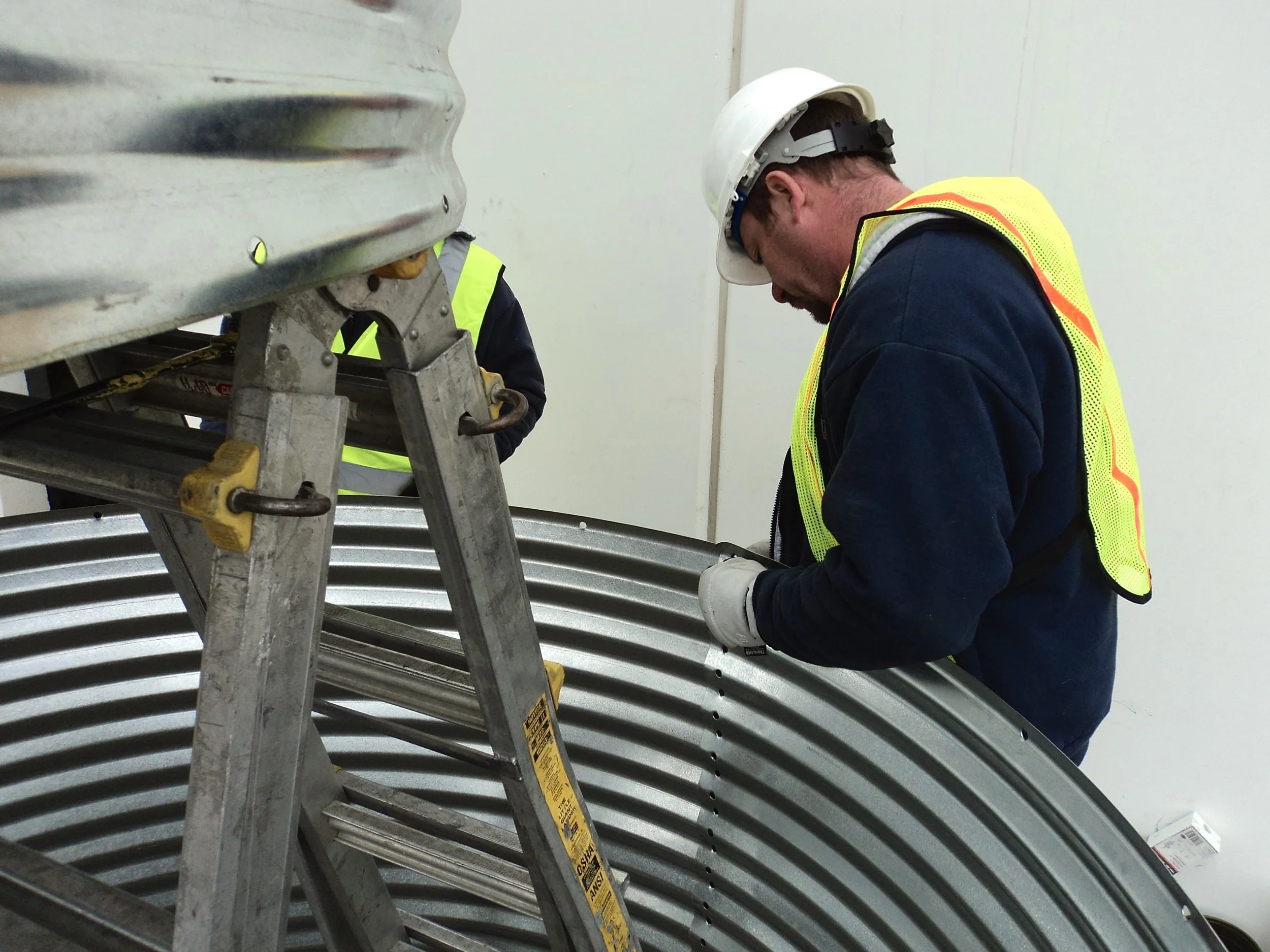 A worker wearing a white hard hat, yellow safety vest, and gloves is inspecting or working on a large metal industrial component or duct, with another worker partially visible in the background.