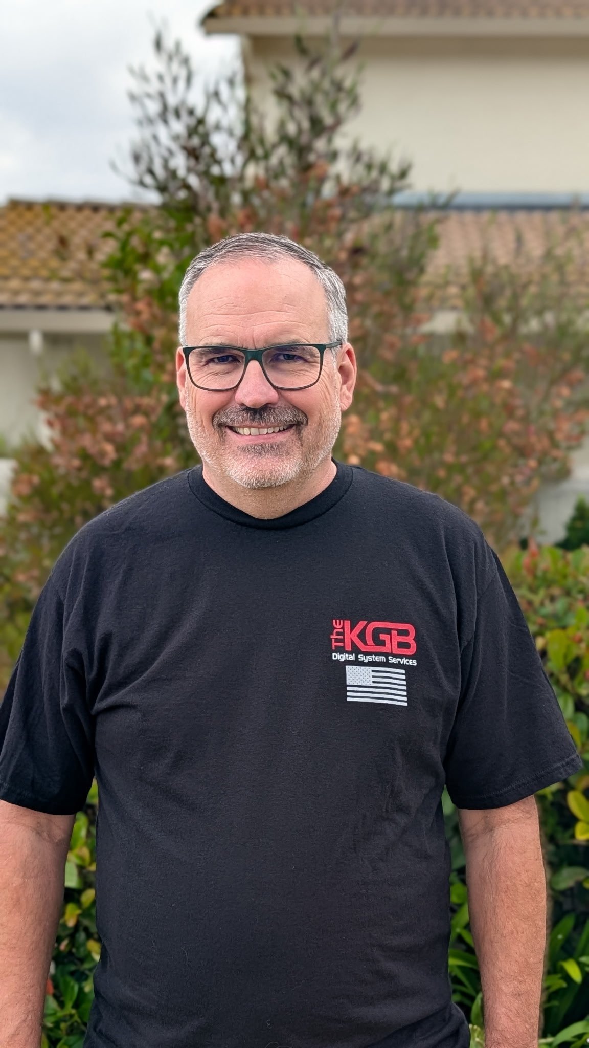 A smiling middle-aged man with gray hair, glasses, and a beard, standing outdoors in front of a garden and house, wearing a black T-shirt with a logo for "The KGB Digital System Services."