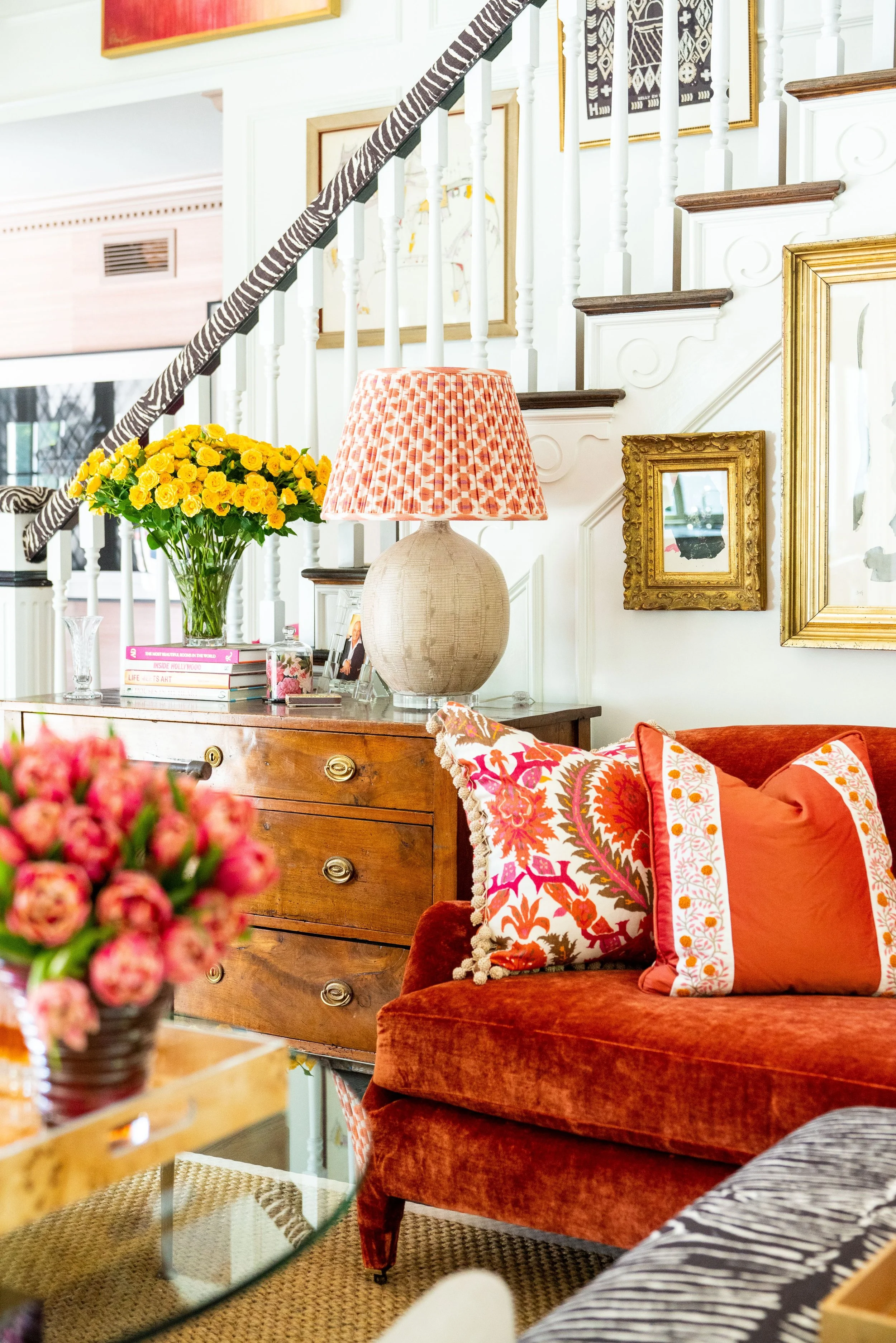 A cozy living room corner decorated with a velvet orange sofa, floral and solid orange throw pillows, a wooden side table with a pink patterned lamp, a vase of yellow flowers, and framed artwork on the wall.