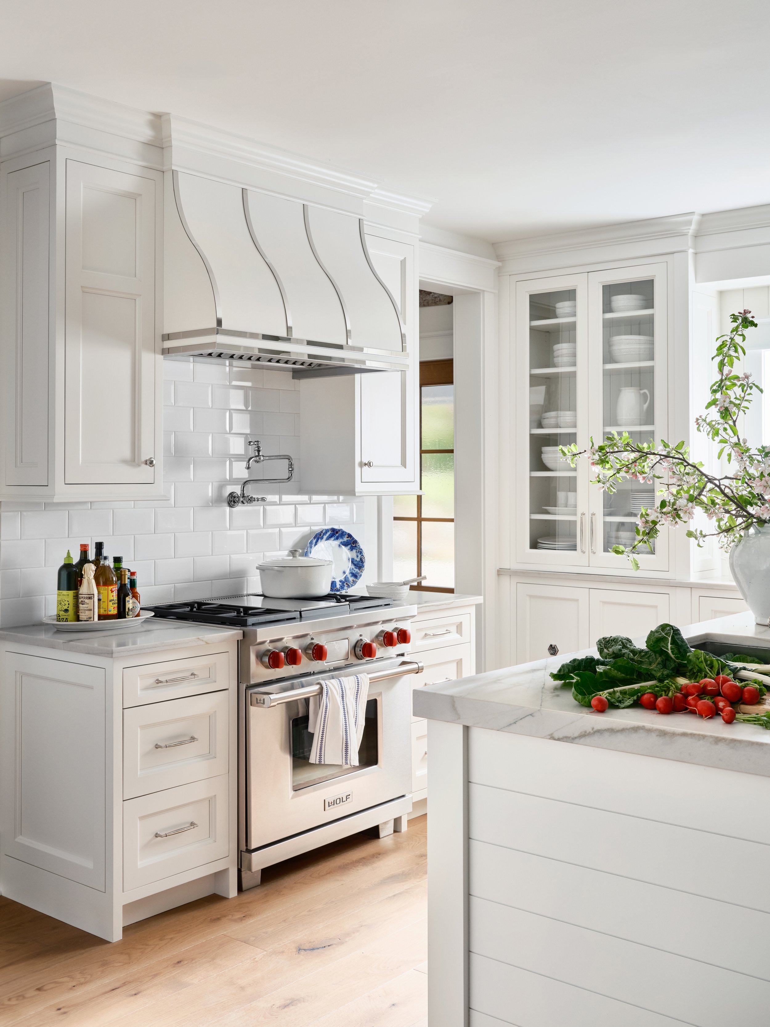 Bright white kitchen with a stove, glass-front cabinets, a vase with cherry blossoms, and fresh vegetables on a marble countertop.