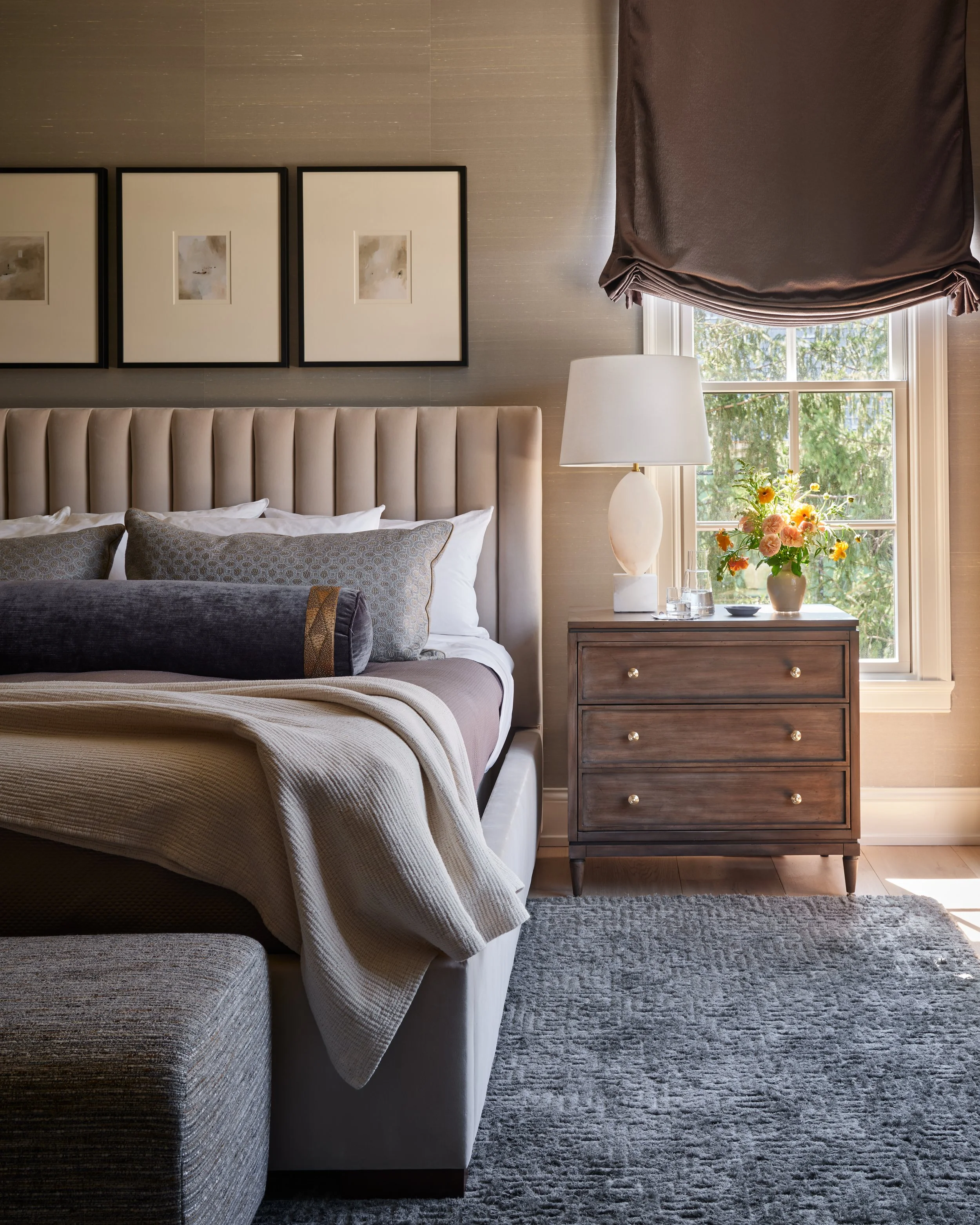 A cozy bedroom with a beige upholstered bed, decorative pillows, a wooden nightstand with a white lamp, a vase of flowers, and a window with a brown curtain, allowing sunlight to illuminate the room.