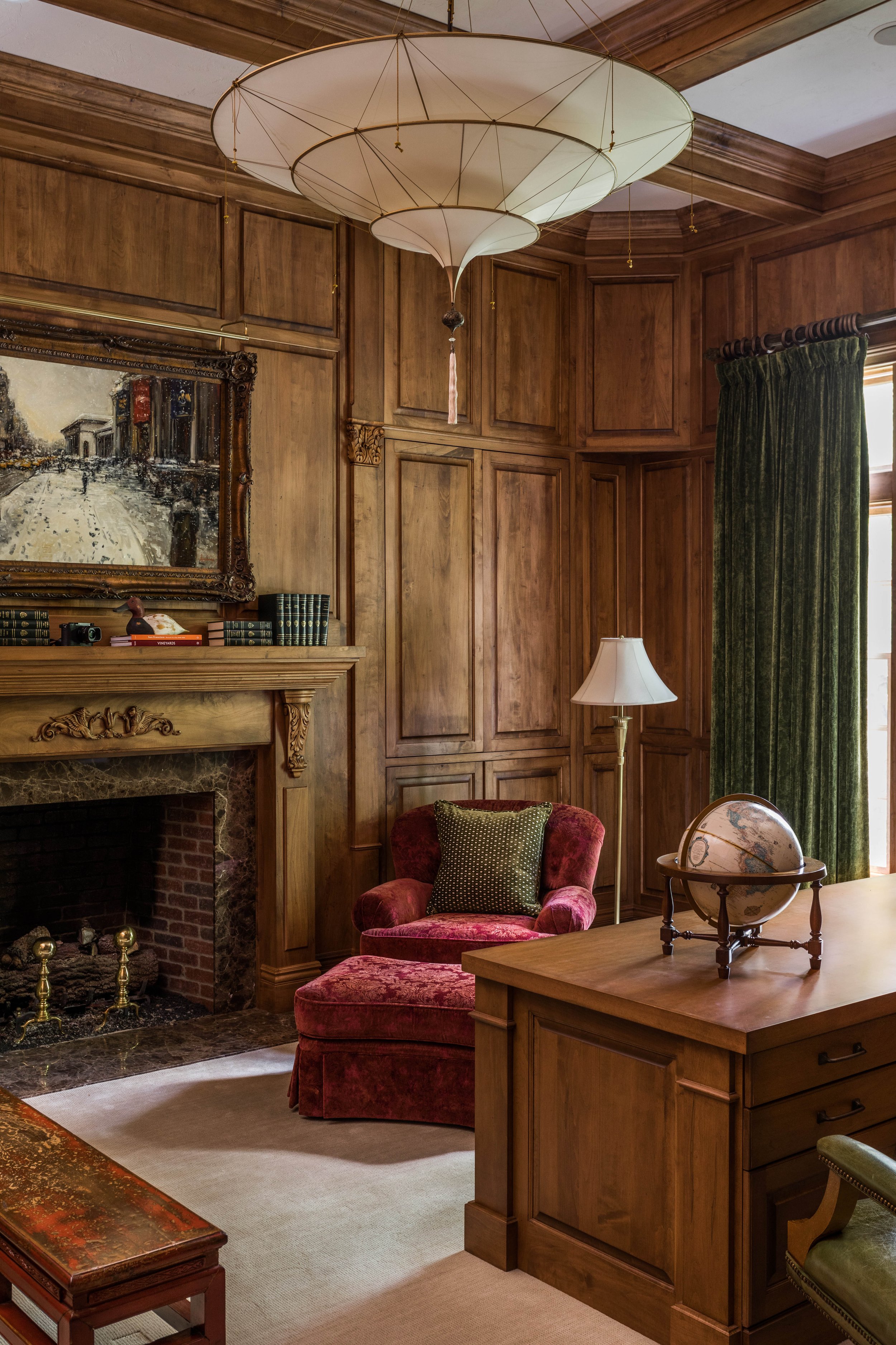 A cozy living room with wooden paneled walls, a fireplace with a marble mantel, an armchair with a green pillow, a globe on a wooden desk, and a large hanging paper lantern light fixture.