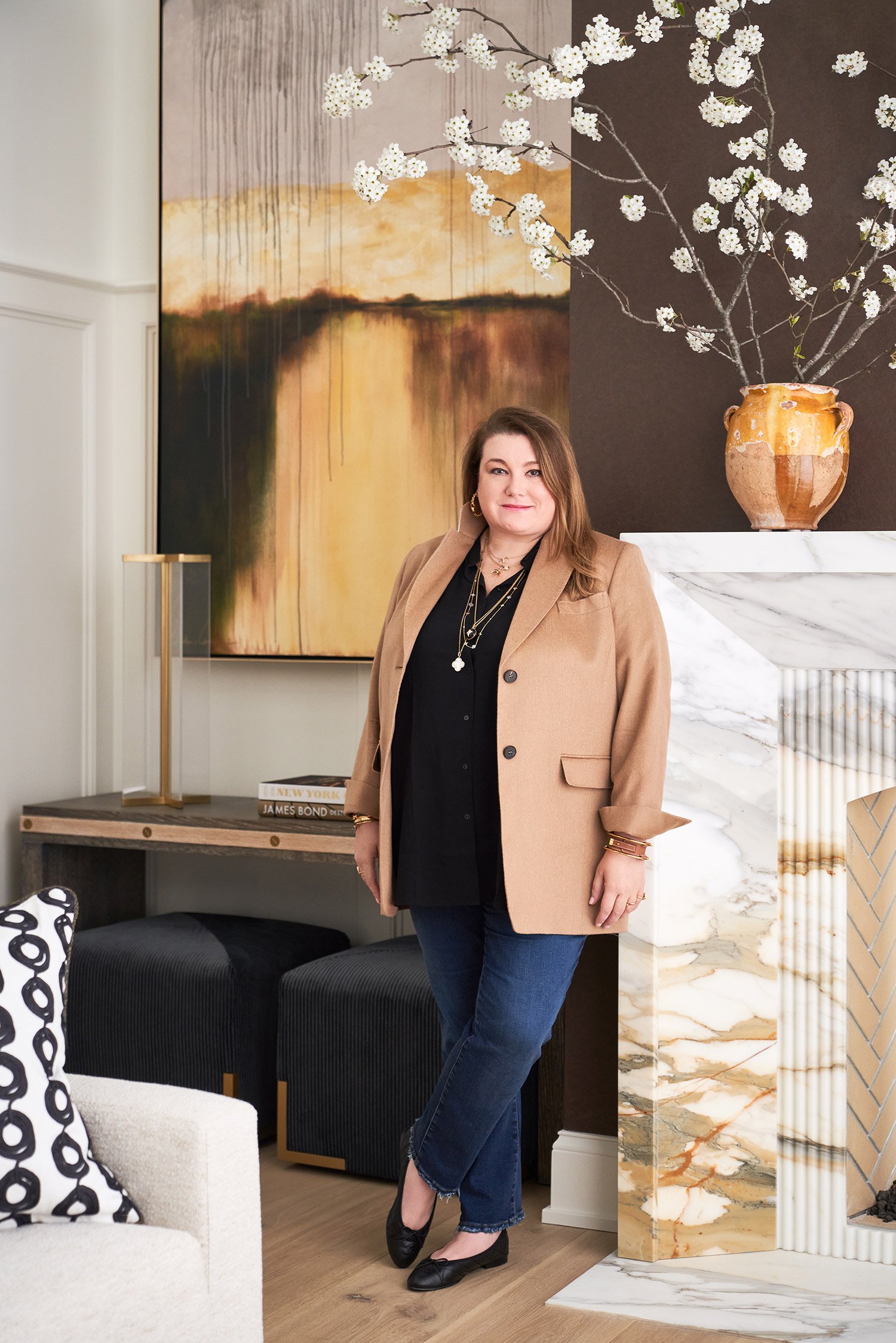A woman with shoulder-length brown hair wearing a tan blazer, black top, blue jeans, and black flats standing next to a marble fireplace in a stylish living room, with abstract art and a large vase with white flowers on the mantel.