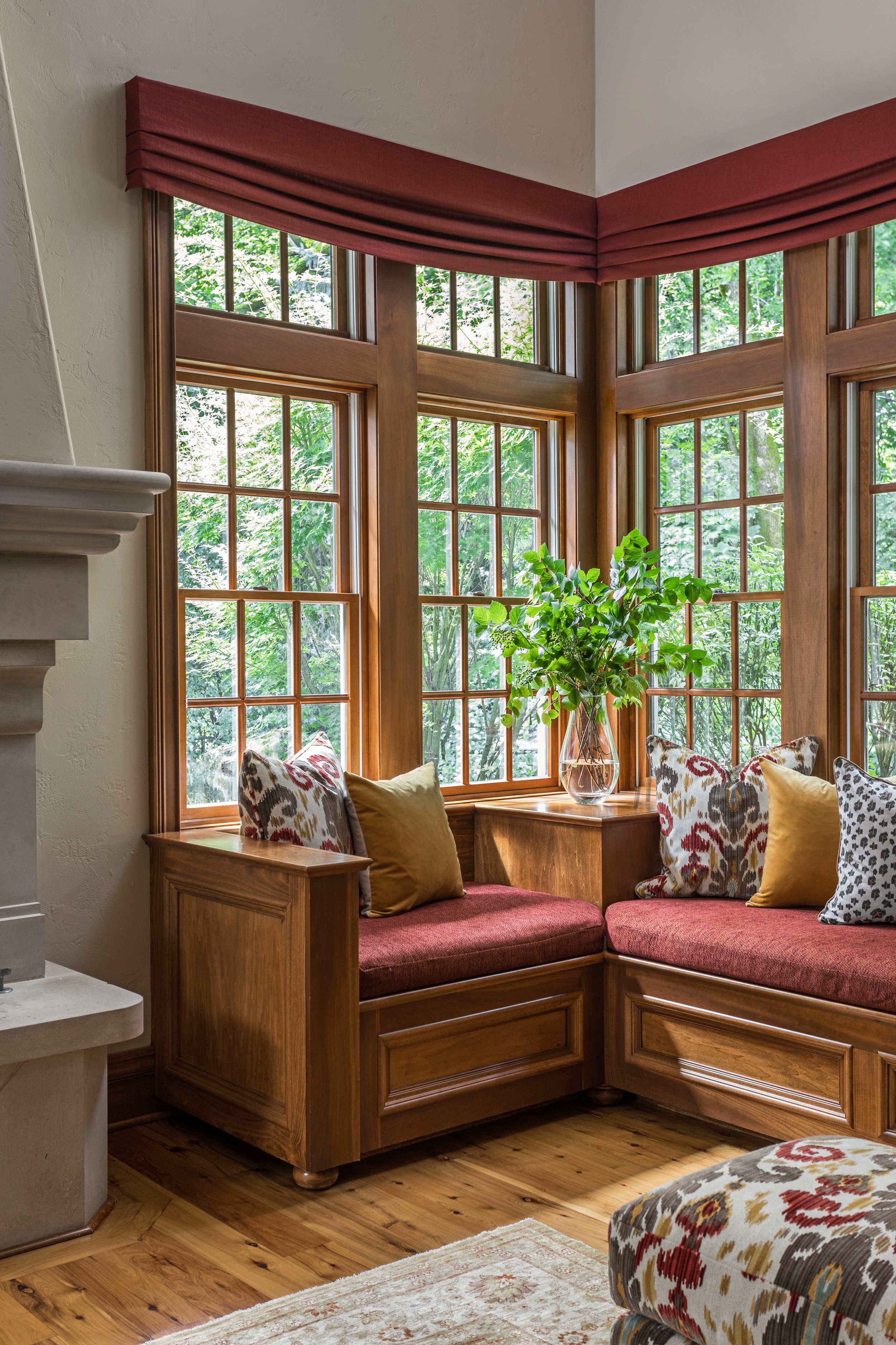 A cozy corner sitting area with a built-in wooden window seat, cushions, and a glass vase with green leafy branches, surrounded by large wooden-framed windows revealing a green outdoor view.
