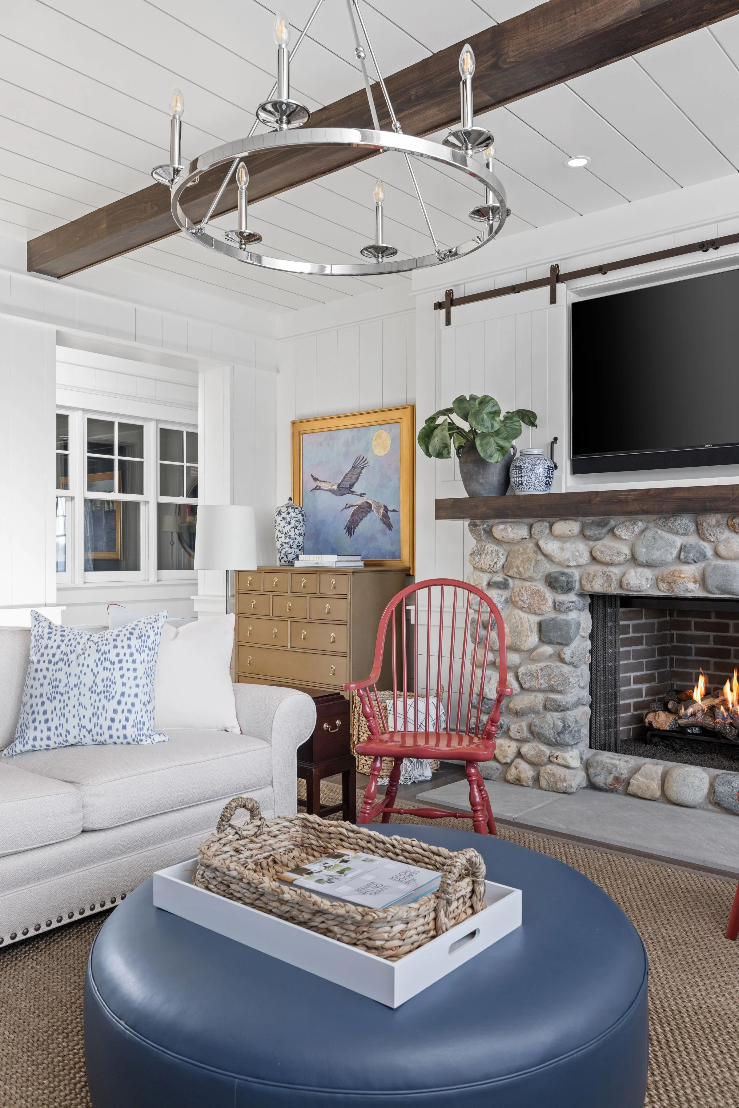 Living room with white sofa, fireplace with stone surround, red wooden chair, black round ottoman with wicker tray, wall art, plants, TV, and chandelier.