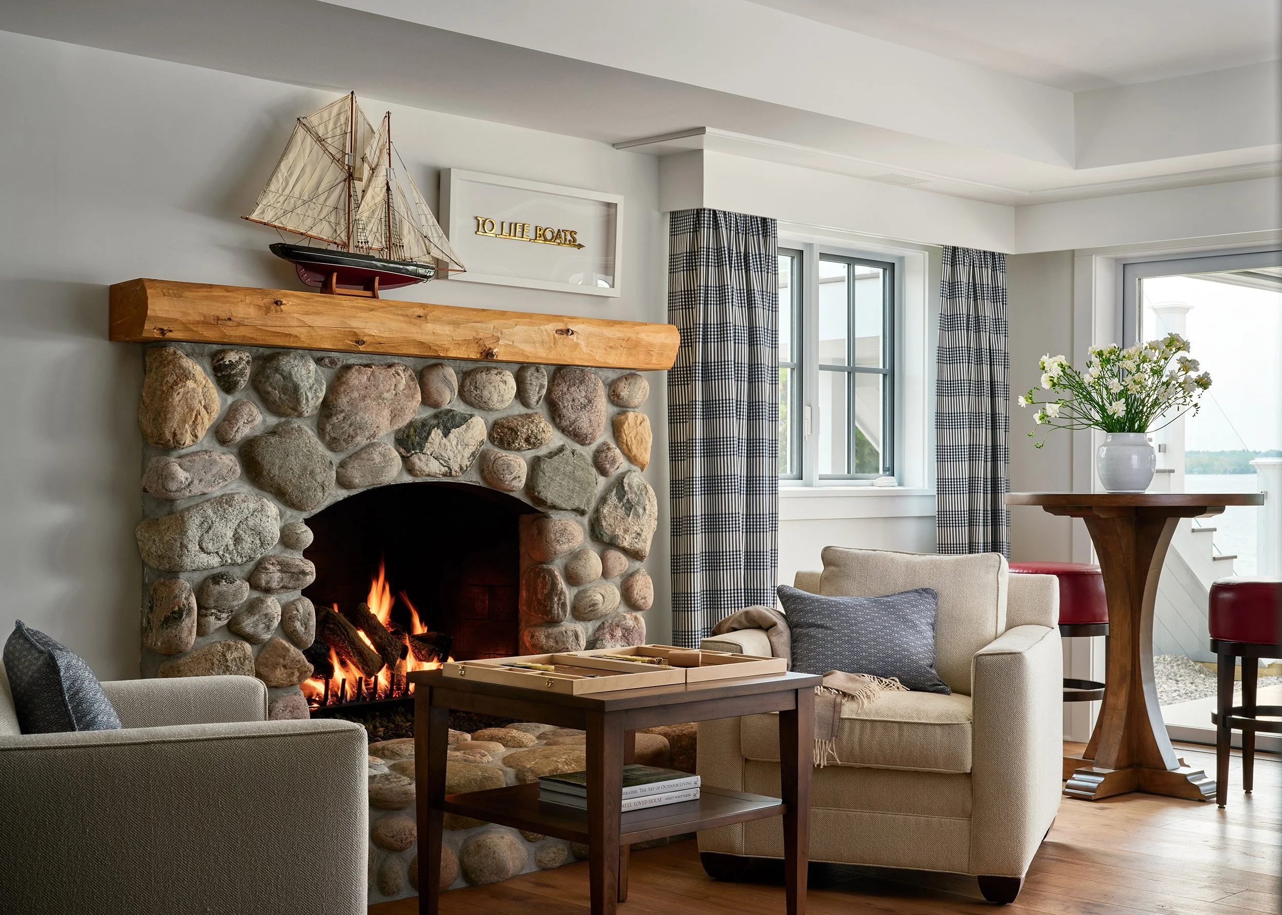 Cozy living room with a stone fireplace, fire burning, beige armchair, coffee table, wooden bar table with flowers, and windows with checkered curtains.