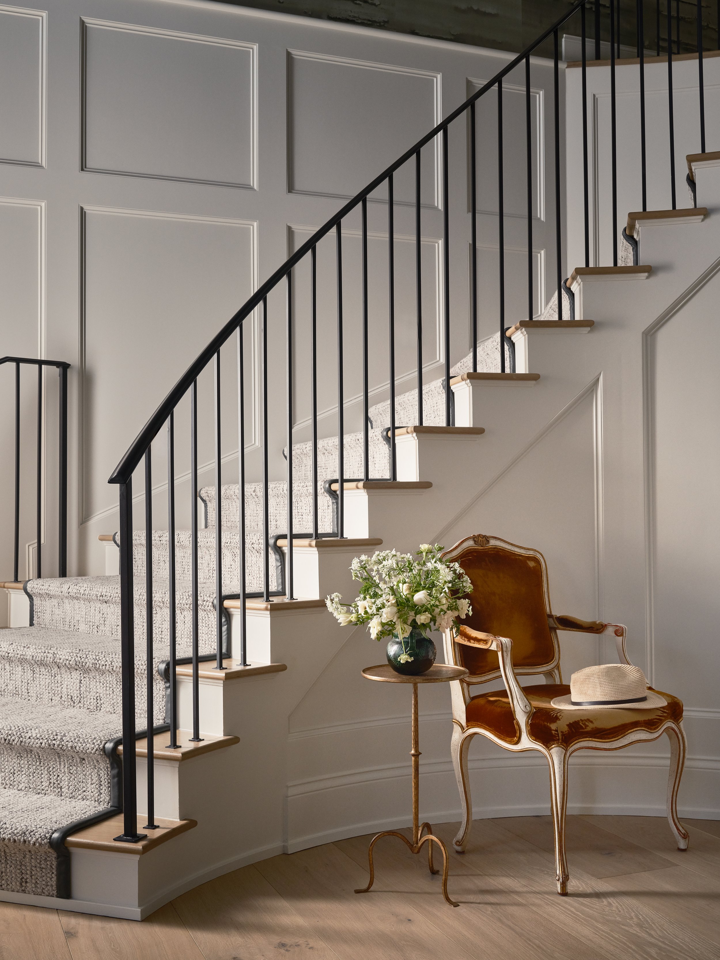 Interior of a home with a curved staircase, beige carpet, white paneled walls, a vintage gold and brown armchair with a straw hat on it, a small wooden side table with a flower vase, and wooden flooring.