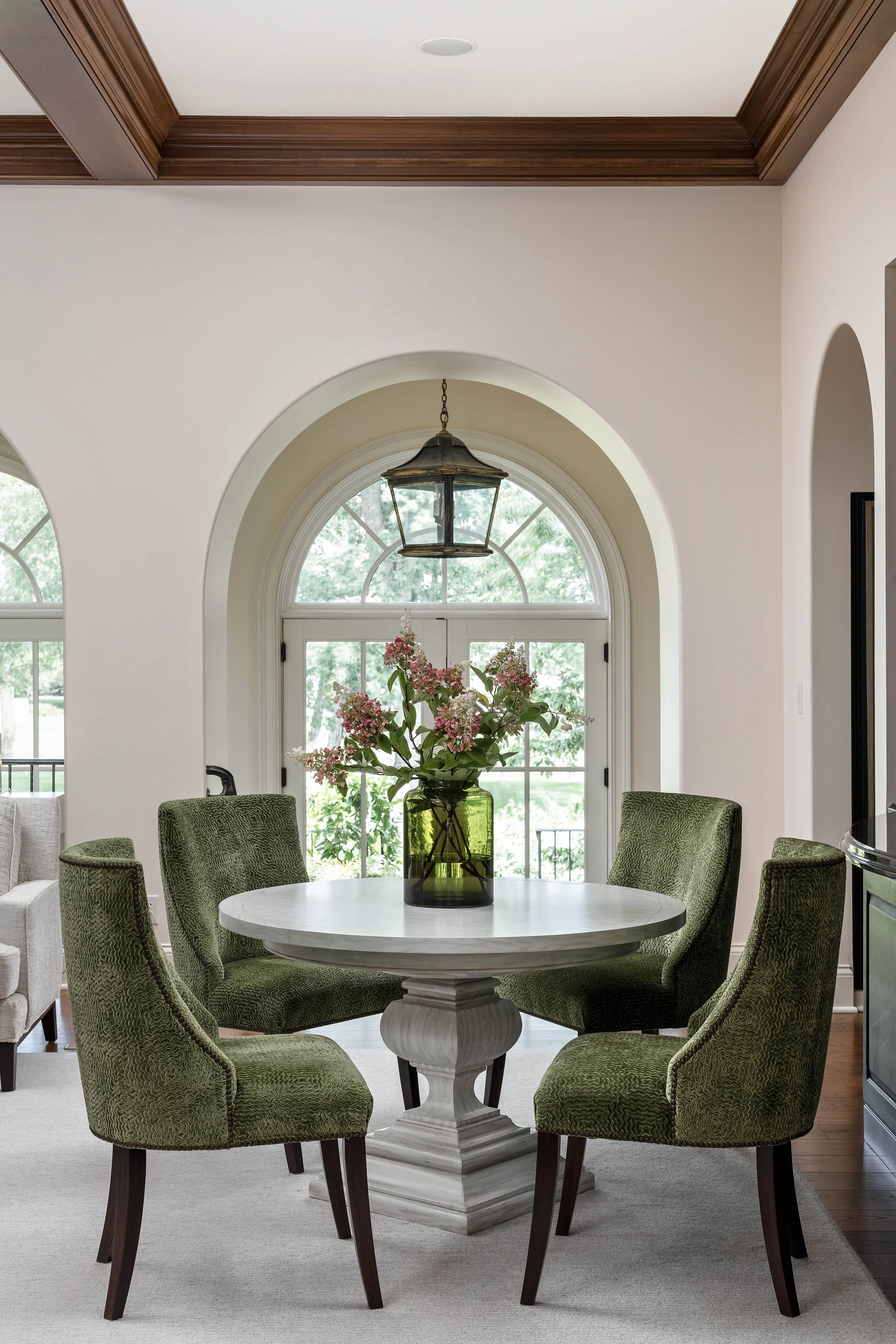 Interior of a dining room with a round white marble table, surrounded by five green upholstered chairs, a black lantern hanging above, and a large window with a semi-circle top in the background, decorated with a vase of pink and white flowers.