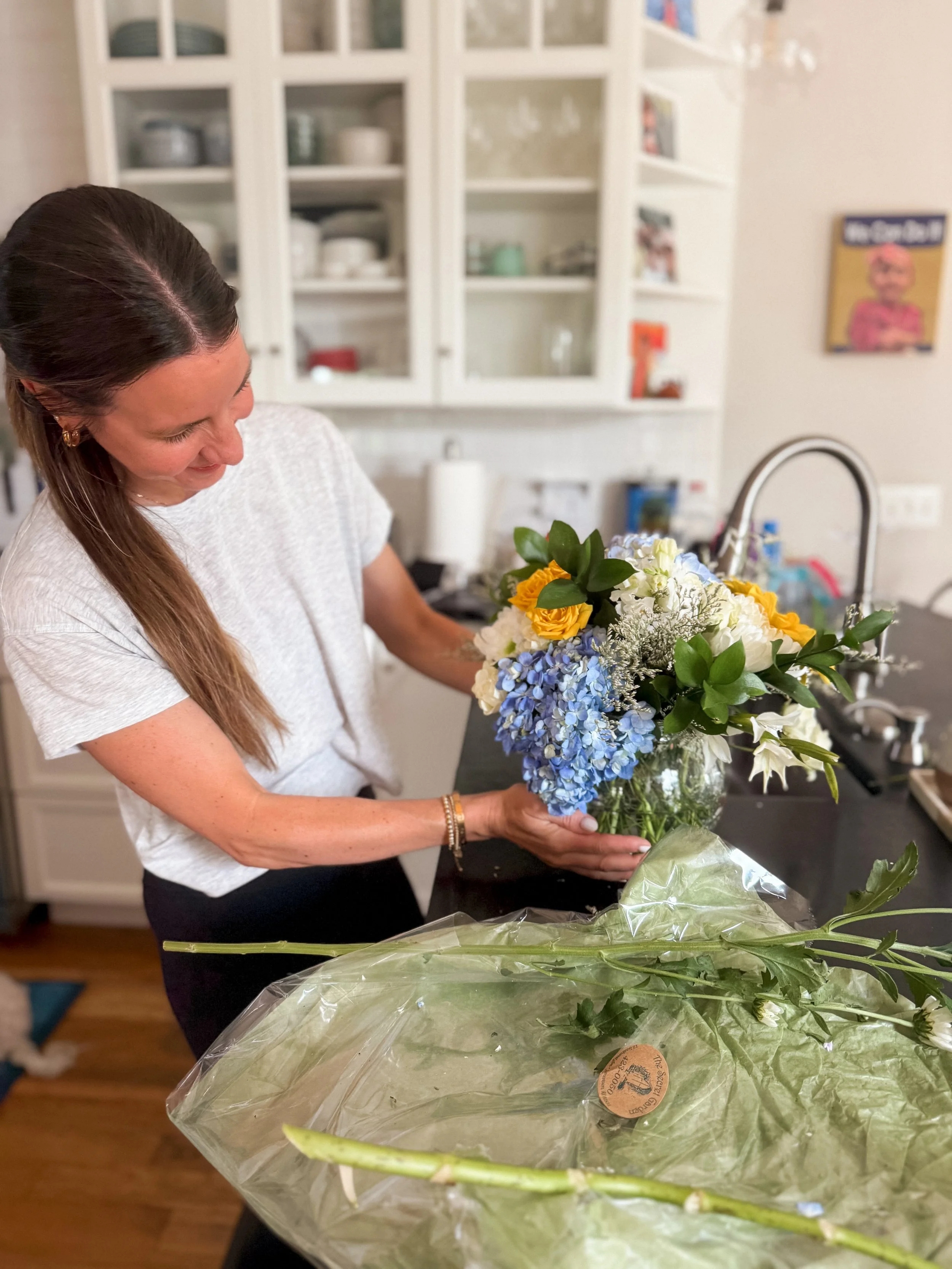 A woman arranging a colorful bouquet of flowers in a kitchen, with white cabinets and a black countertop.