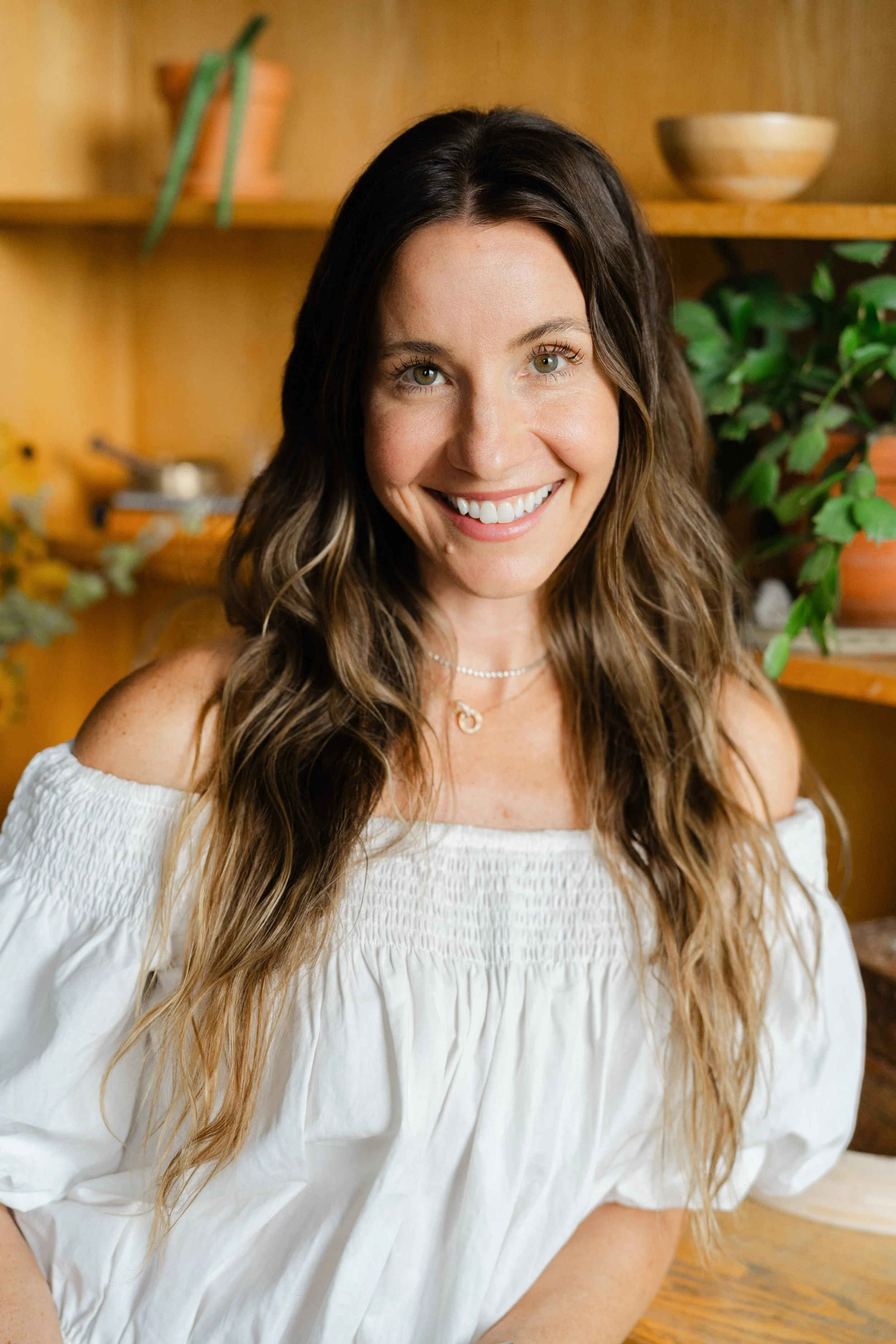 A woman with long, wavy brown hair and light skin smiling in front of a wooden shelf with plants and pottery.