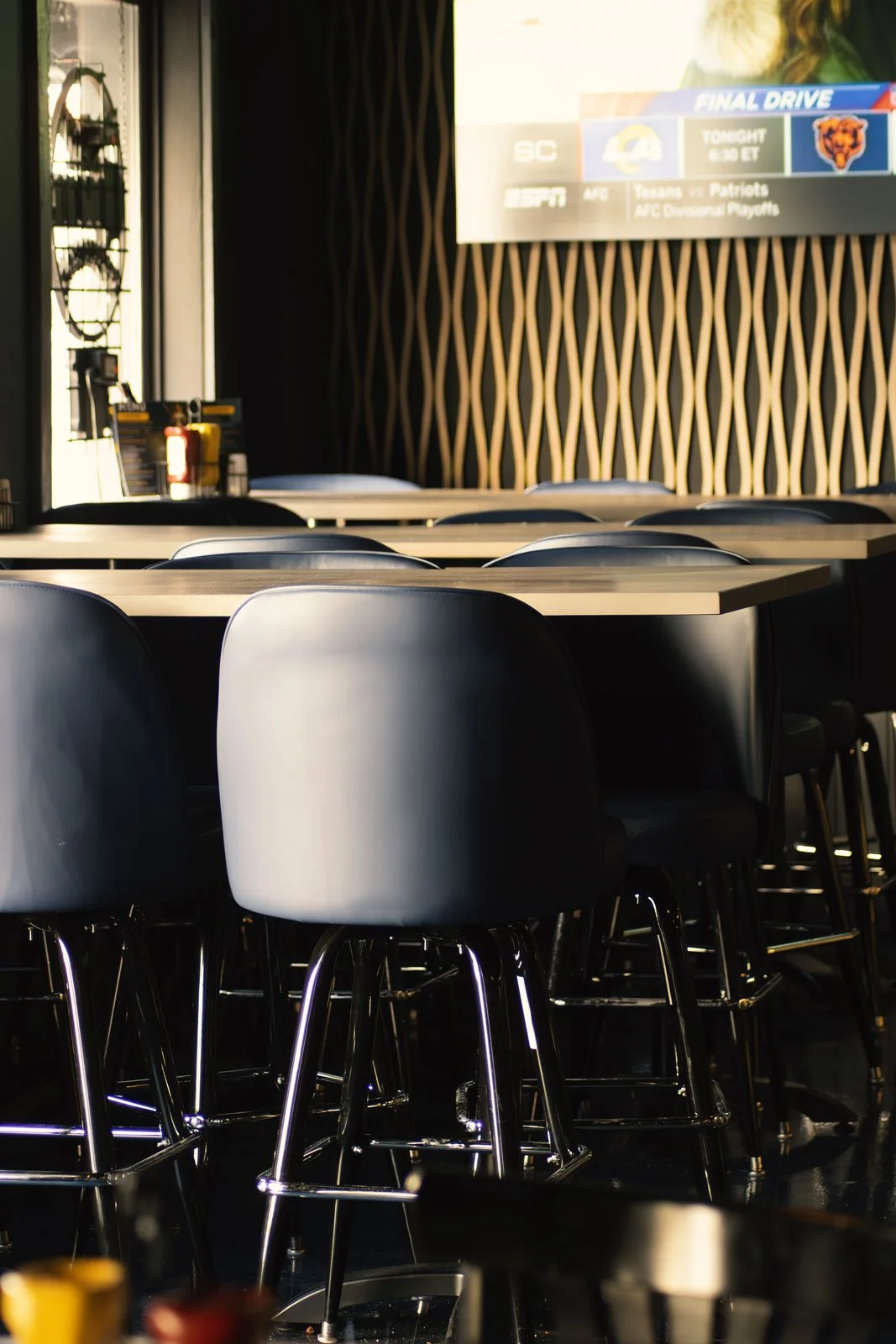 Empty bar stools and tables inside a restaurant or bar, with a large screen displaying sports and TV ratings in the background.