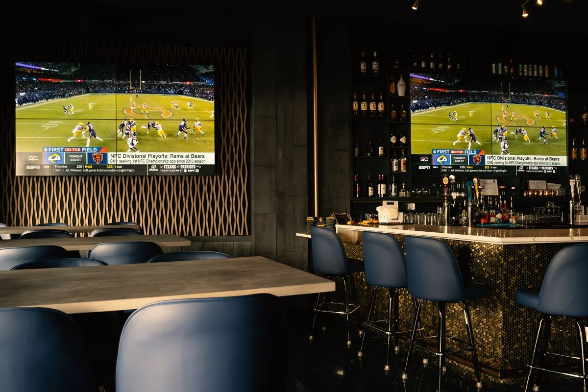 Two large flat-screen TVs displaying a football game are mounted on the wall in a bar with a dark interior. There are several blue barstools lined up at the bar counter, which holds bottles of alcohol and glasses. Several tables and chairs are arranged in the foreground.