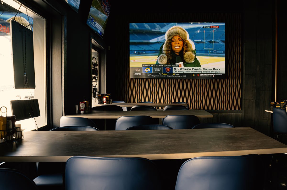Chic Sports Bar, restaurant with dark wood tables and black chairs, large windows on the left, and a large flat-screen TV on the back wall showing a news broadcast about an NFL game with a woman reporting outside the stadium.