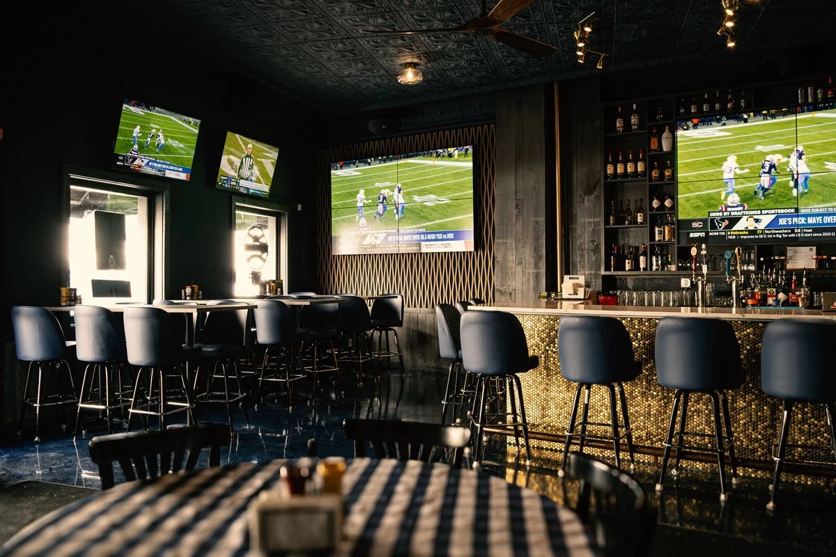 Indoor sports bar with multiple flat-screen TVs showing football games, illuminated bar area with black barstools, and a dark ceiling with fans, wood-paneled walls, and large windows.
