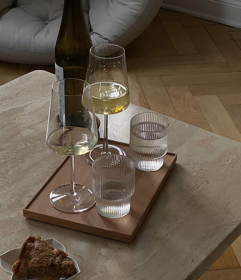 A tray with two glasses of white wine, two glasses of water, and a wine bottle on a marble table, with a piece of pie on a small plate in the foreground and a cozy chair in the background.