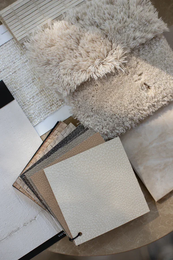 A collection of fabric or wallpaper samples in neutral tones and textures, with two plush, furry throw blankets in light beige and gray on a round table.
