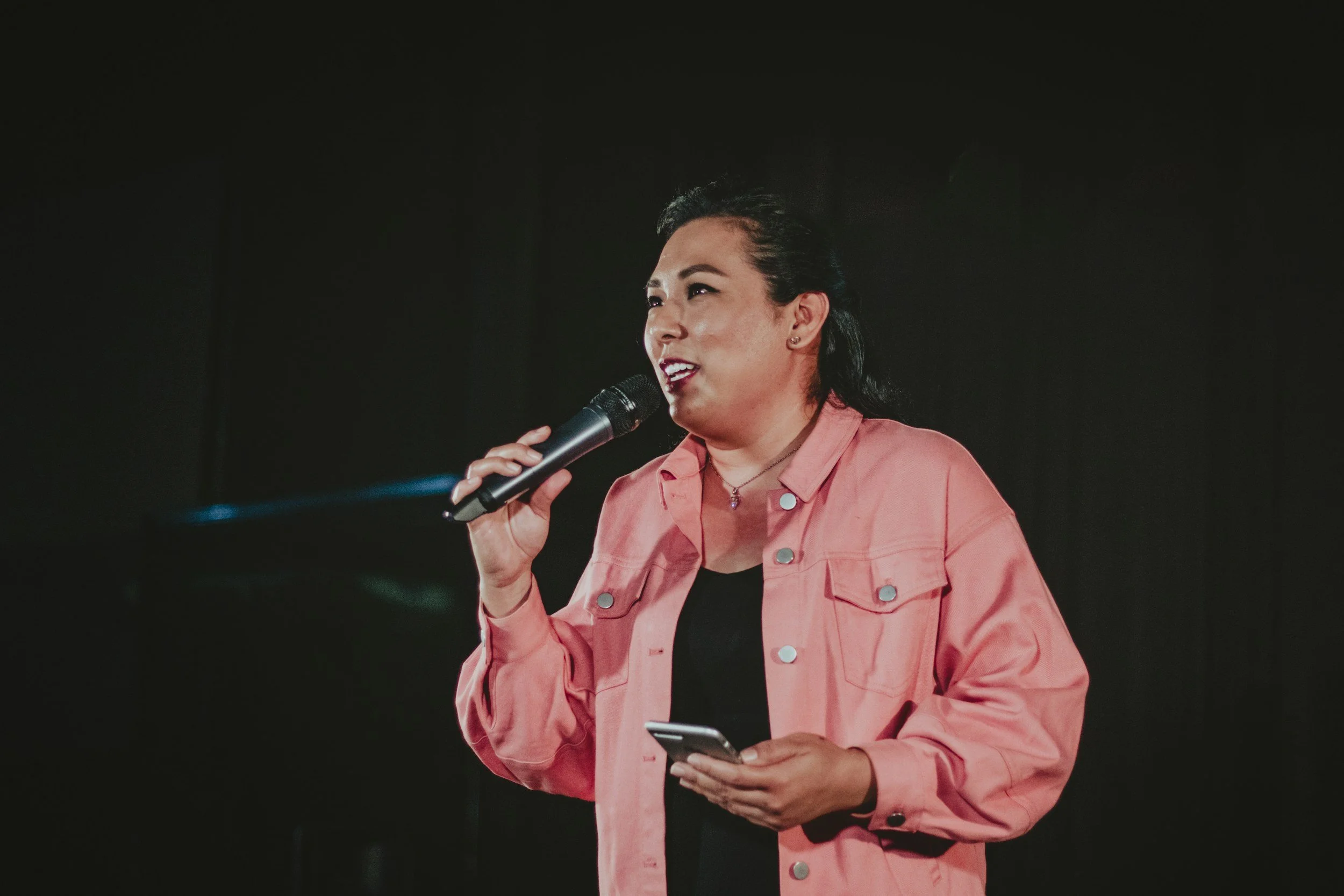 Woman holding a microphone and smartphone, speaking on stage against a dark background.