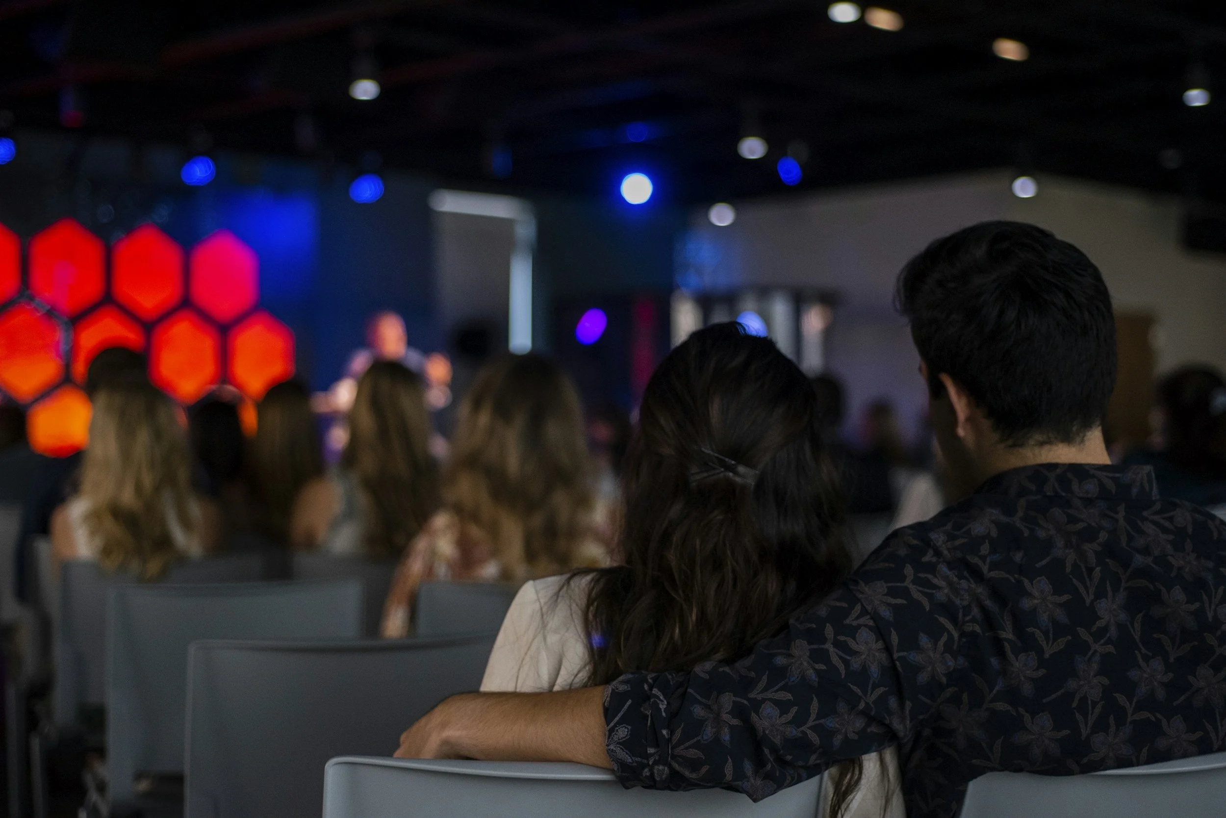 Couple sitting closely together at a concert or event, with their arms around each other, watching a speaker or performer on stage in a dimly lit room.
