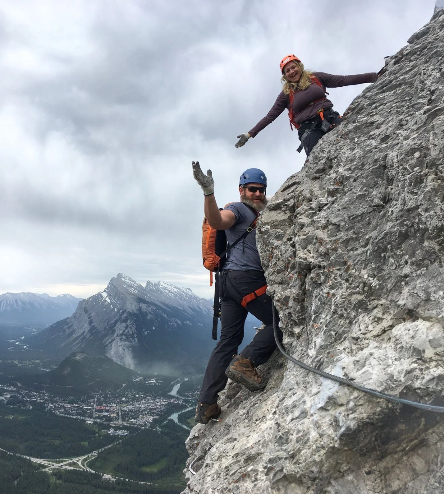 Two climbers, a woman and a man, on a steep mountain face with snow-capped peaks and a valley in the background. They are wearing helmets and climbing gear, and are waving at the camera.