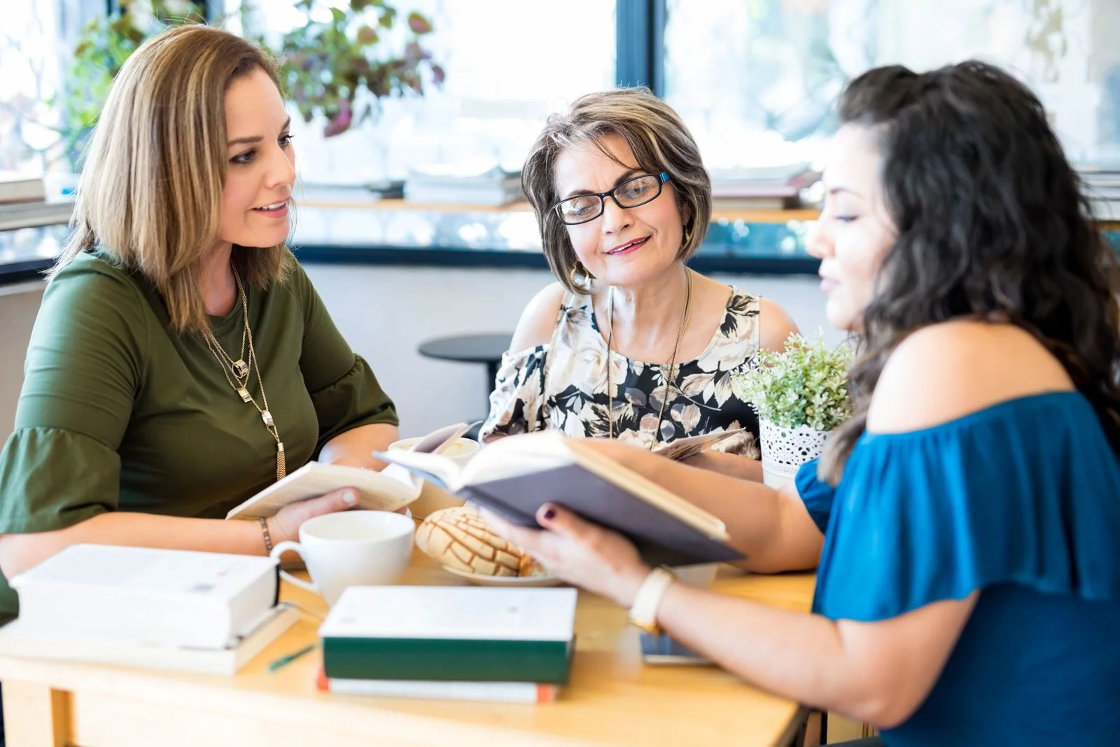 Three women sitting at a table in a bright room, looking at books and discussing, with notebooks and a potted plant on the table.