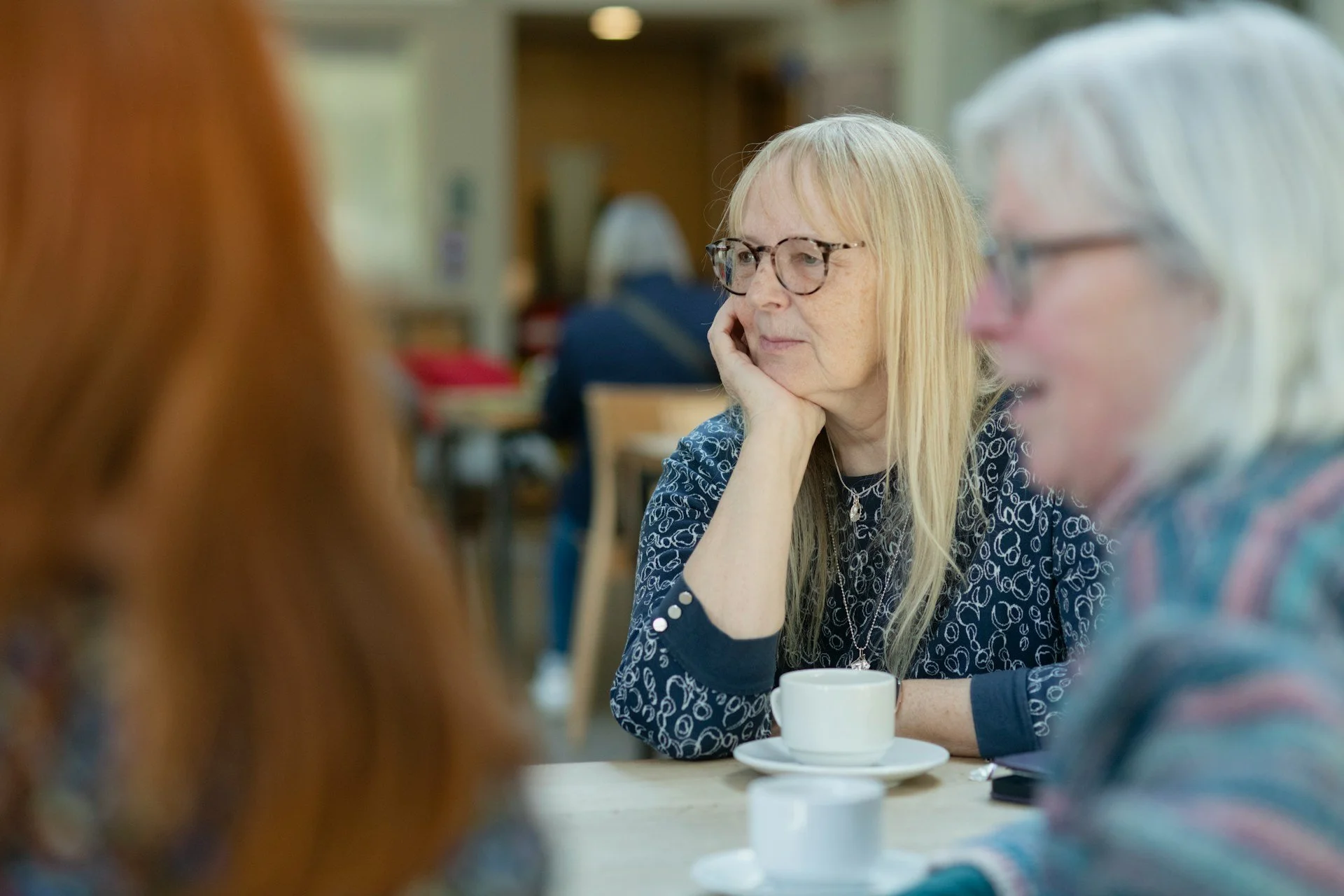 Three women sitting at a table in a casual indoor setting, engaged in conversation. One woman with blonde hair and glasses appears to be listening thoughtfully, while the other two, one with red hair and the other with white hair, are partially visible.