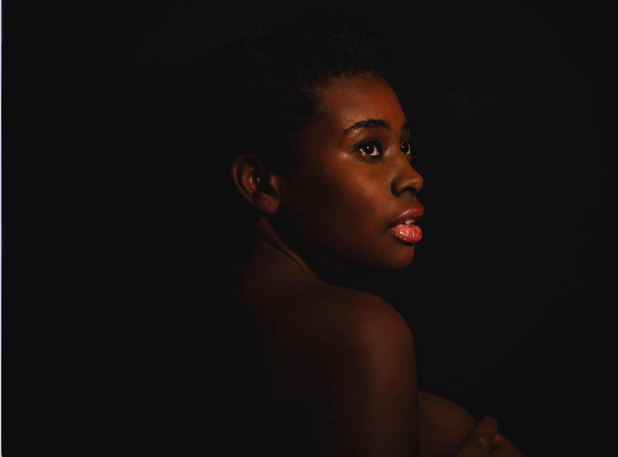 Portrait of a young Black woman with short natural hair against a dark background, with soft lighting highlighting her face, shoulder, and lips.