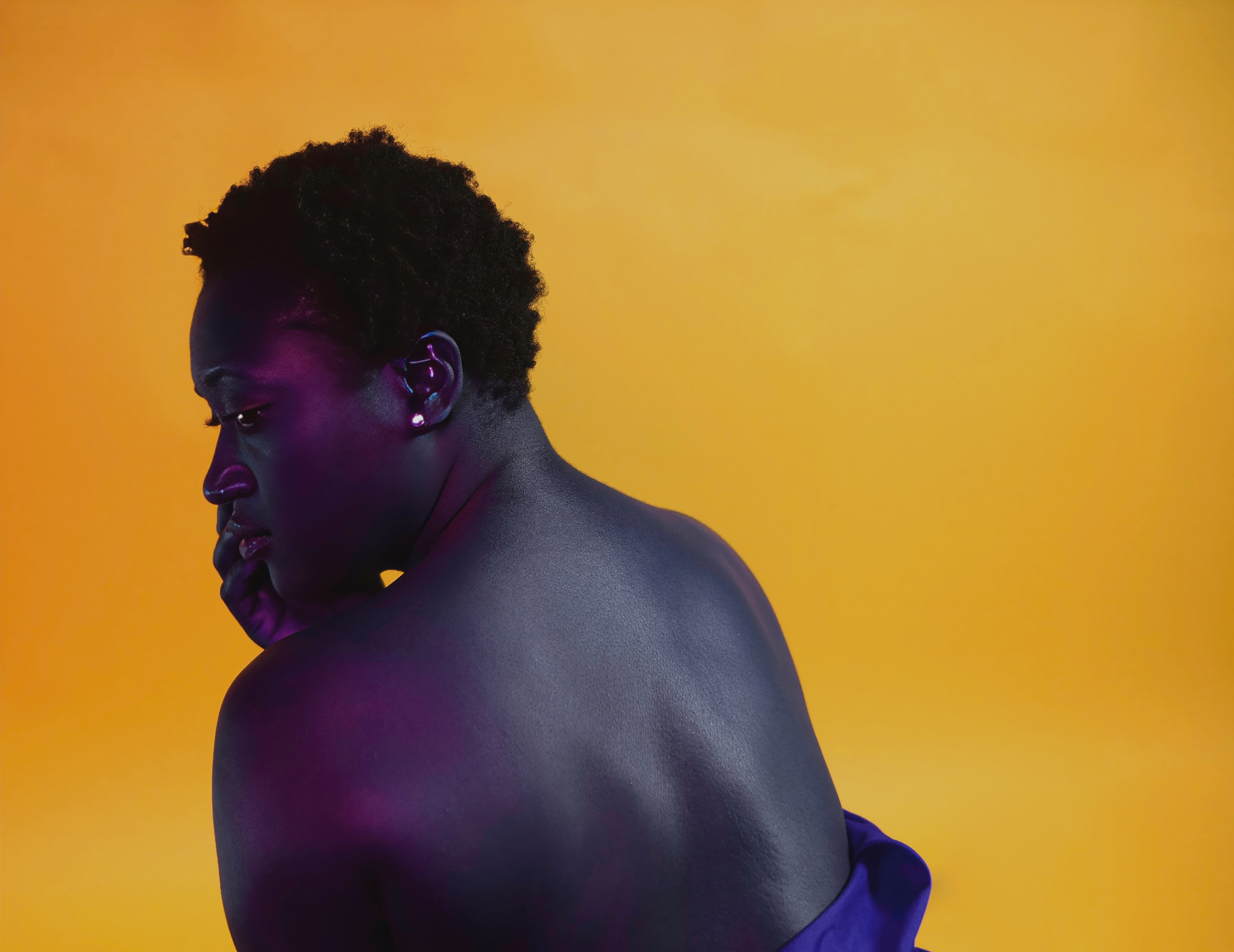 A woman with short, curly hair and diamond earrings is posing against a yellow background. She is looking down and resting her chin on her hand, with her back partially turned to the camera.
