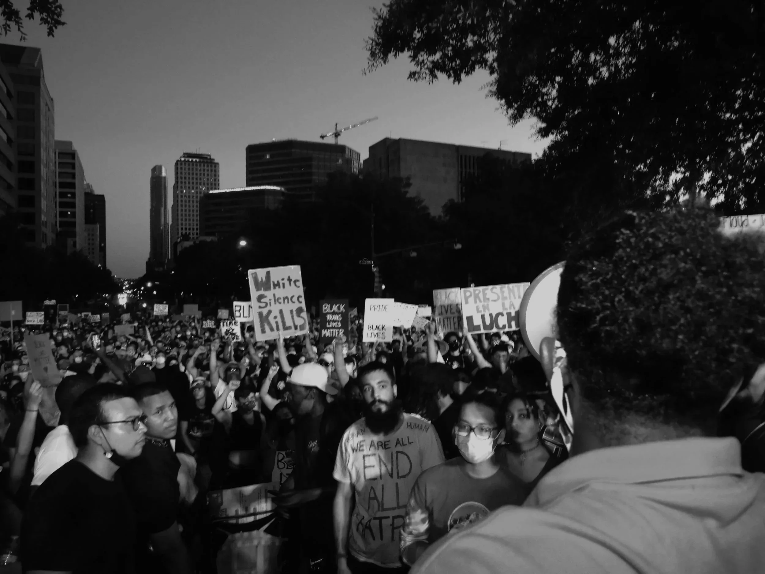 Crowd of protesters holding signs during a demonstration in an urban area at dusk, with tall buildings and city skyline in the background.