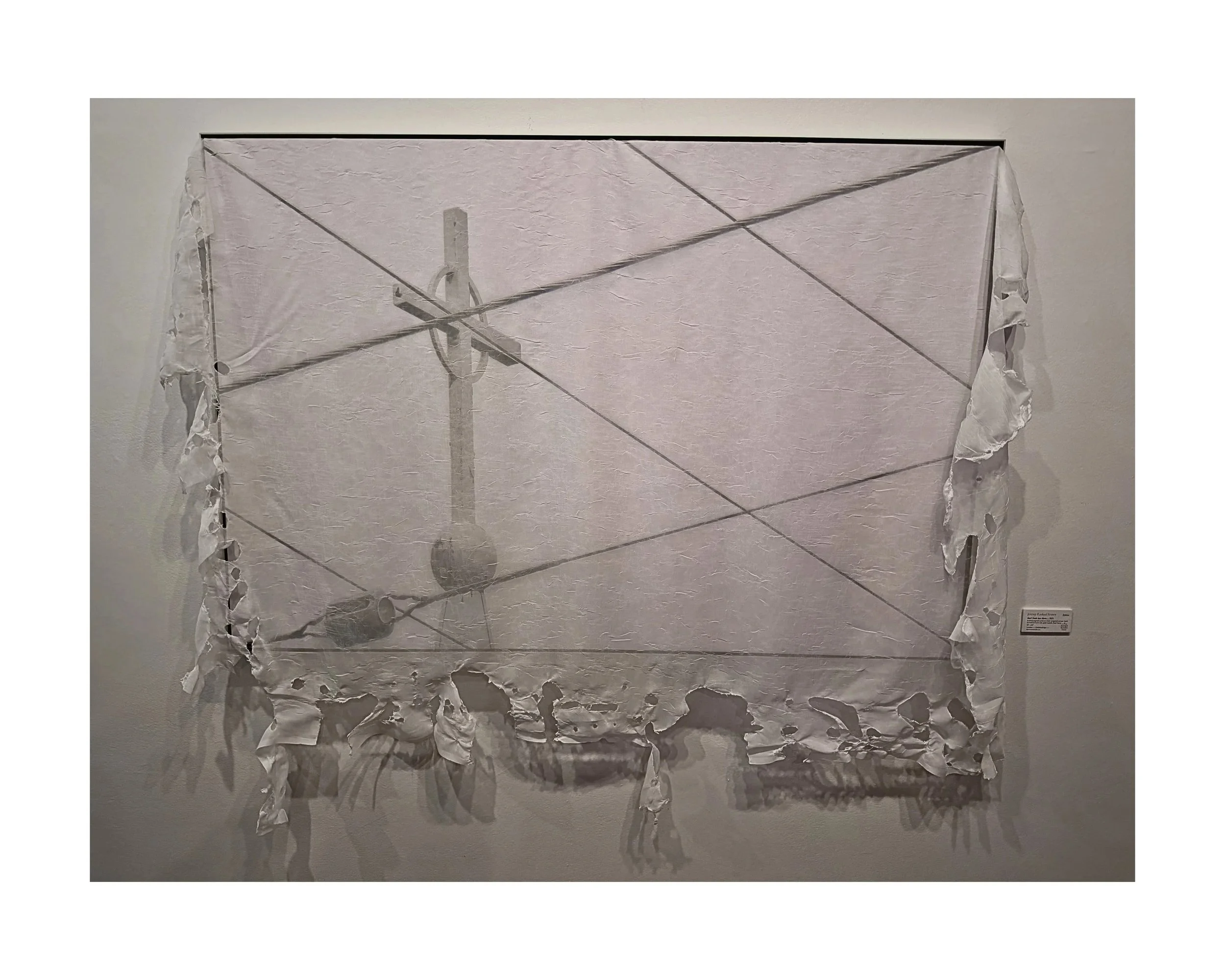 Art piece resembling a torn, white, tattered flag with a black silhouette of a pole and wires, mounted on a white wall.