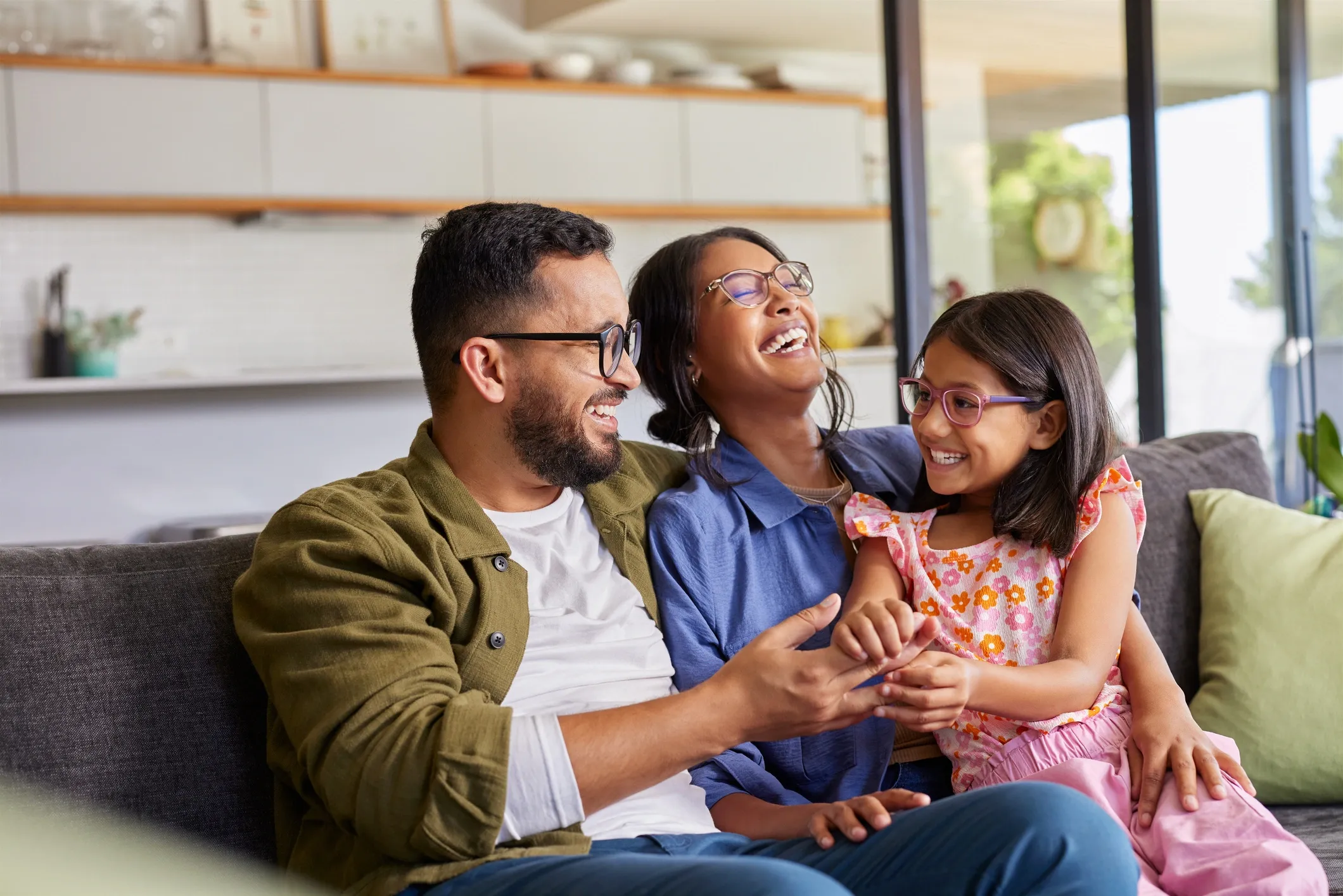 Family of three sitting on a sofa, smiling and laughing together in a living room with large windows and a kitchen in the background.