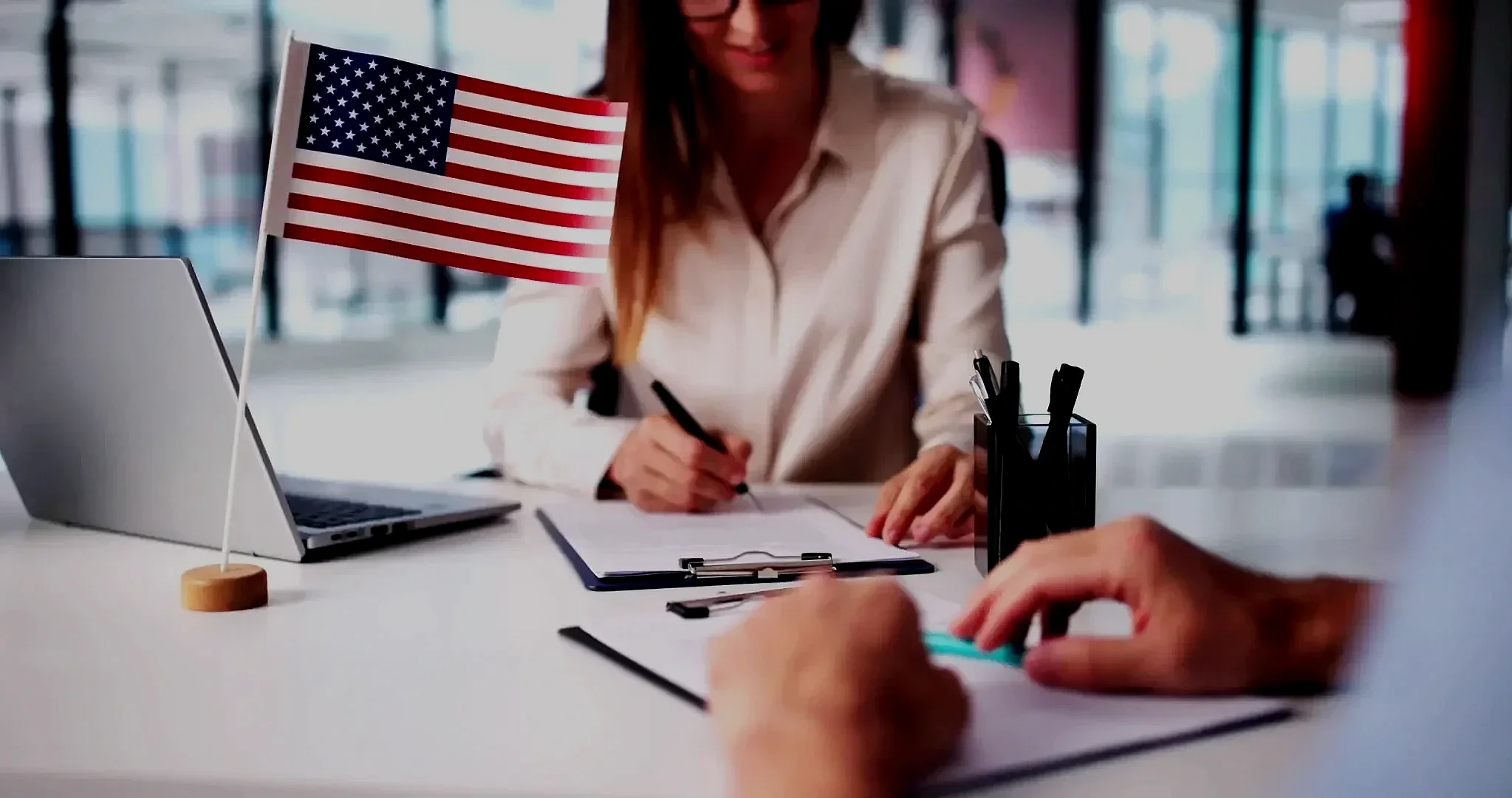 A woman sitting at a desk, filling out paperwork, with a small American flag on the desk, a laptop, and a pen holder.