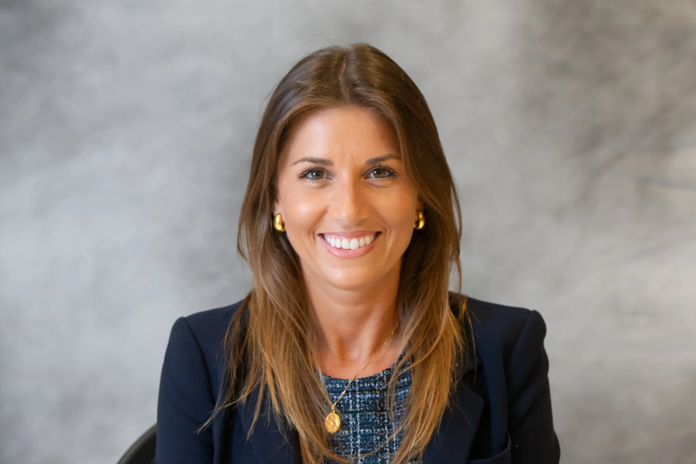Smiling woman with brown hair wearing a dark blazer and gold jewelry against a gray background.