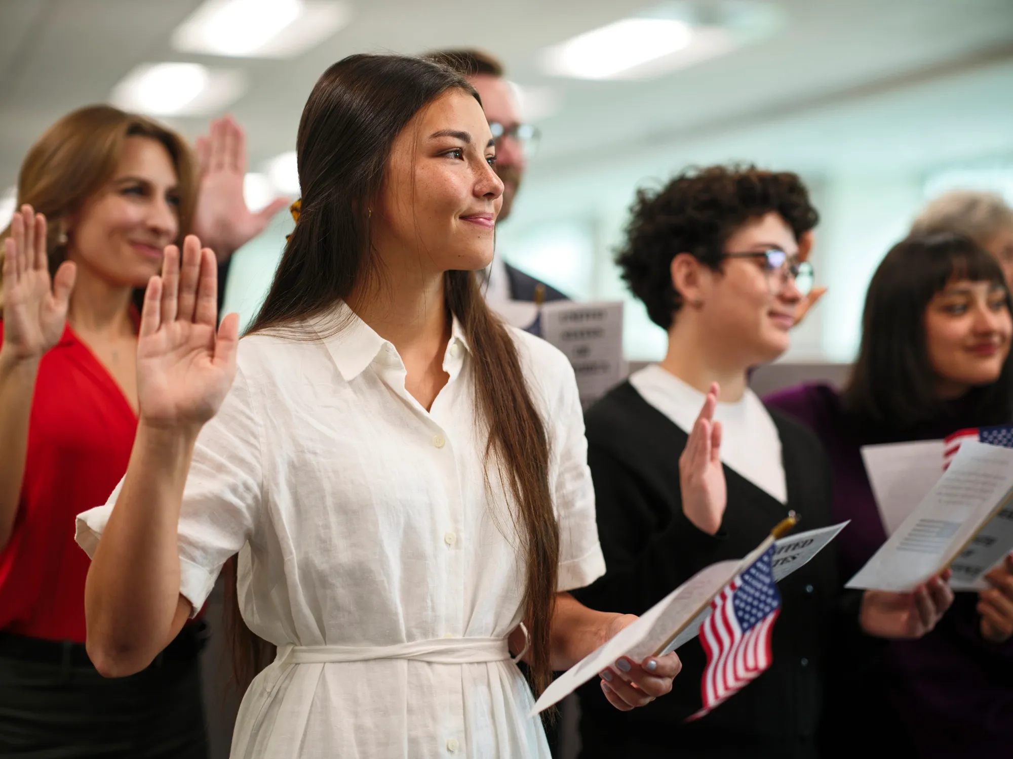 Group of diverse people raising their right hands during an oath-taking ceremony, with some holding American flags and pamphlets.